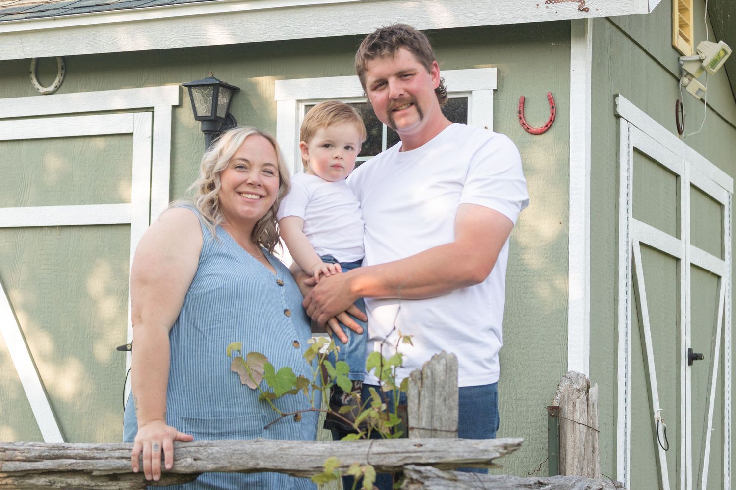 Smiling family of three posing by rustic fence near green shed.