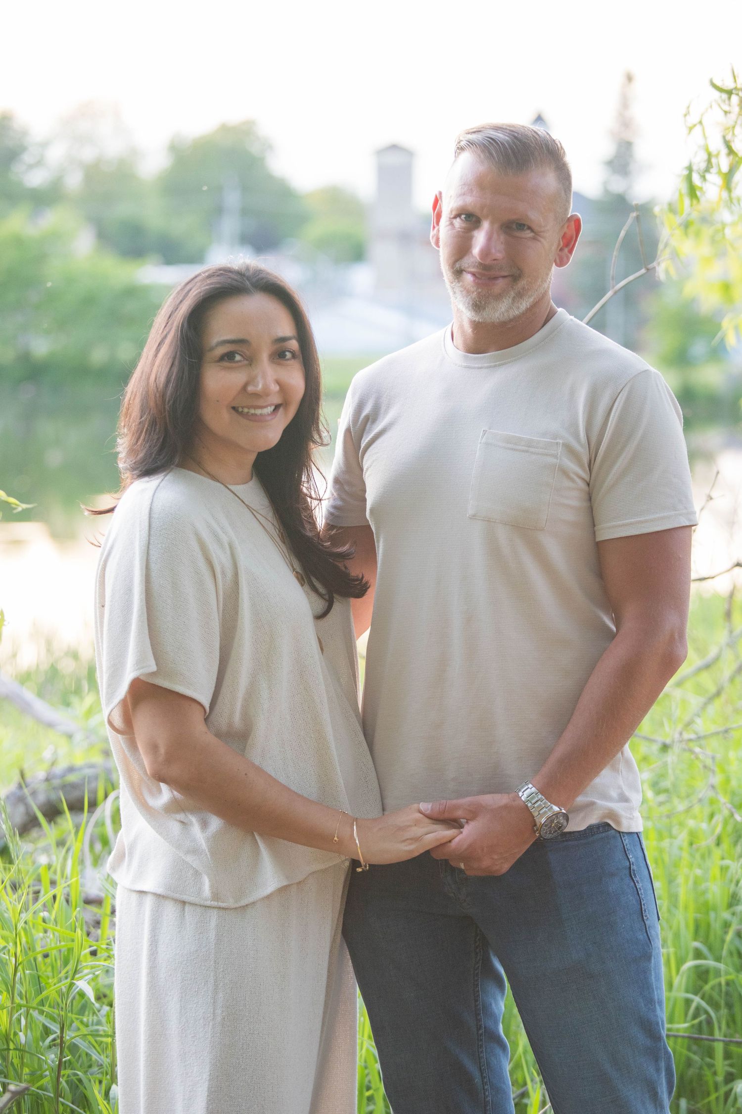 Smiling couple holding hands outdoors near a river, wearing matching beige outfits in a lush green setting.