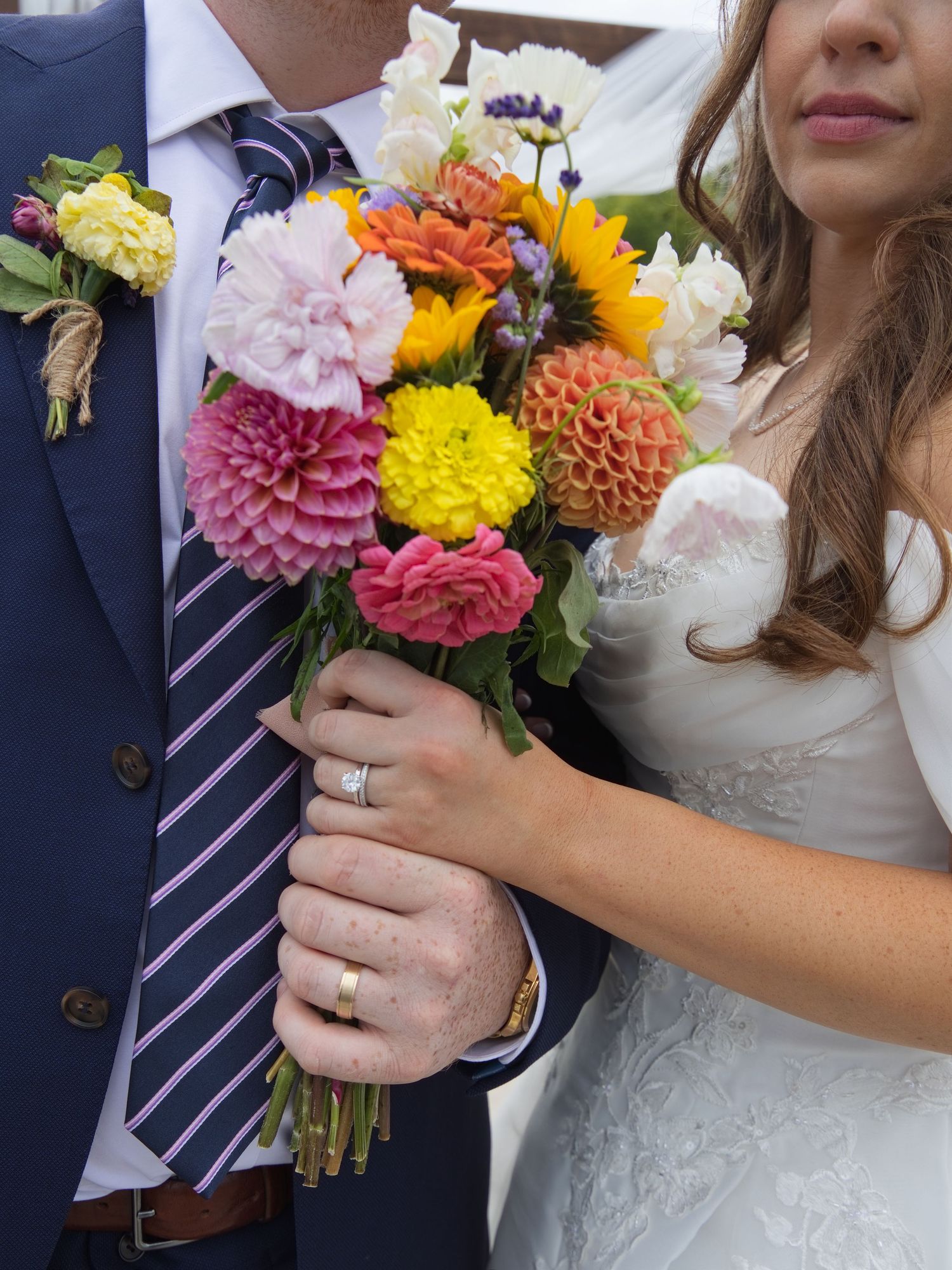 Bride and groom holding a vibrant wedding bouquet with dahlias, sunflowers, and mixed blooms, both wearing gold wedding bands.