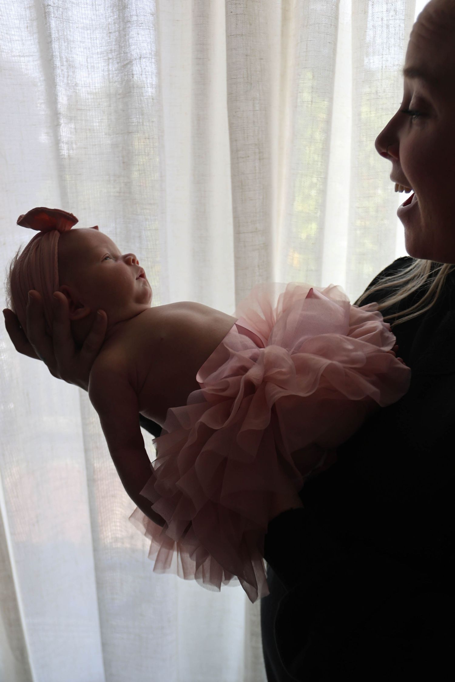 Mother holding newborn baby girl in pink tutu dress near white curtains.