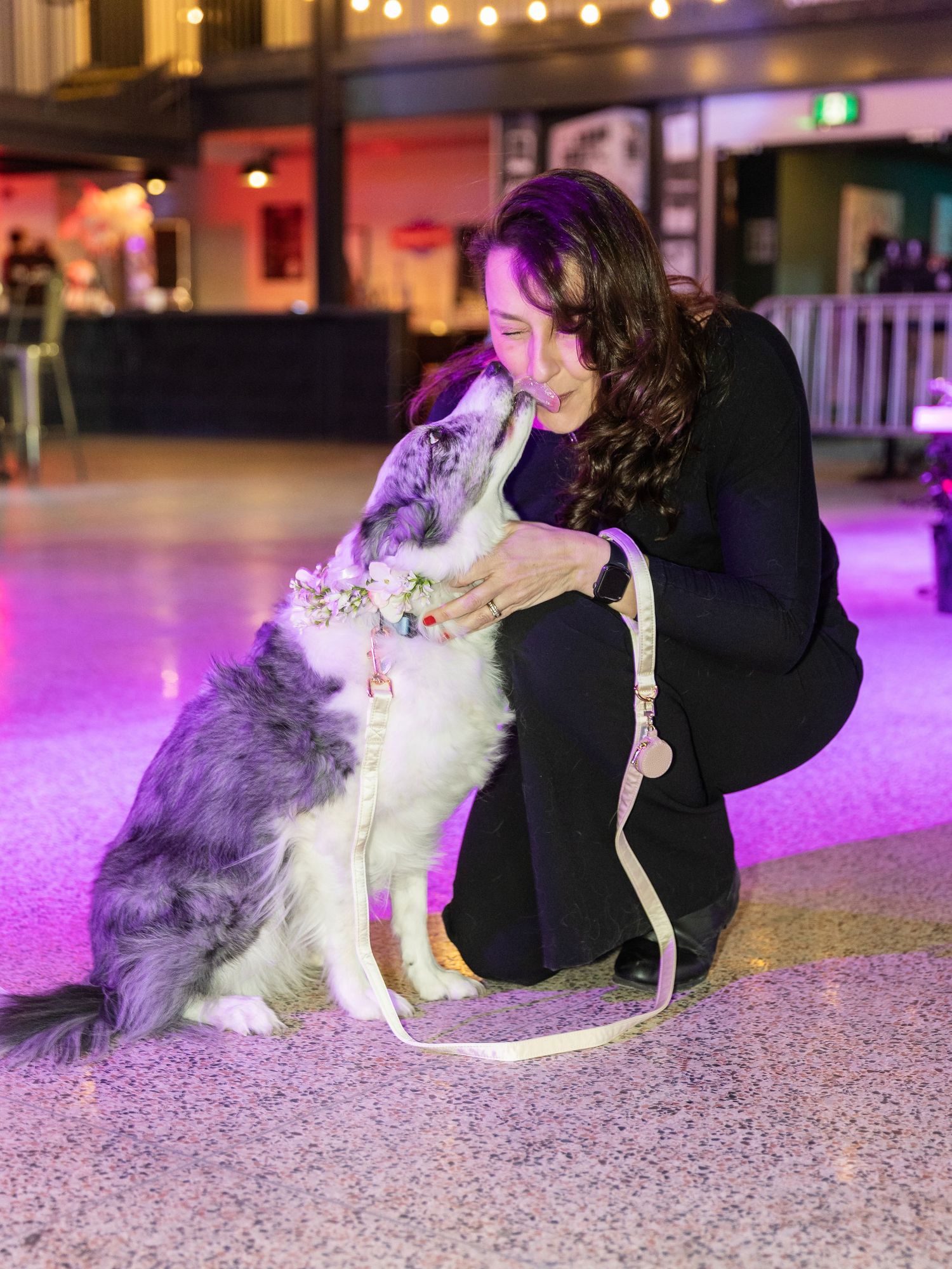 Woman laughing as fluffy dog with flower crown gives kisses on vibrant pink lit venue floor.