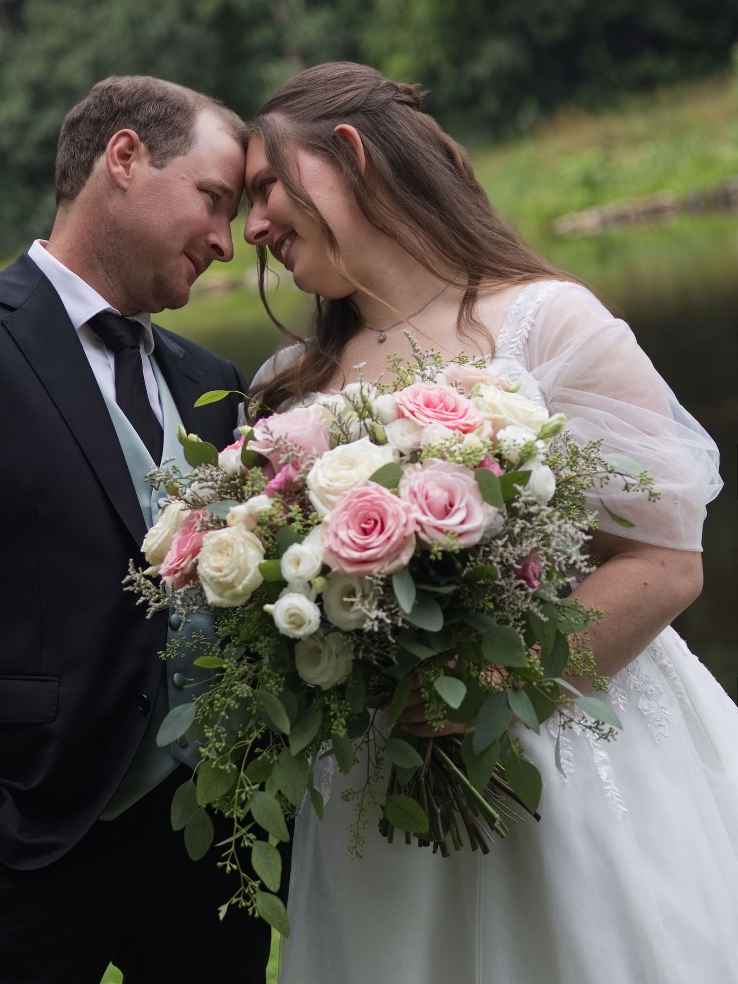 Bride and groom touching foreheads tenderly, bride holding pink and white rose bouquet with greenery, outdoor wedding portrait.
