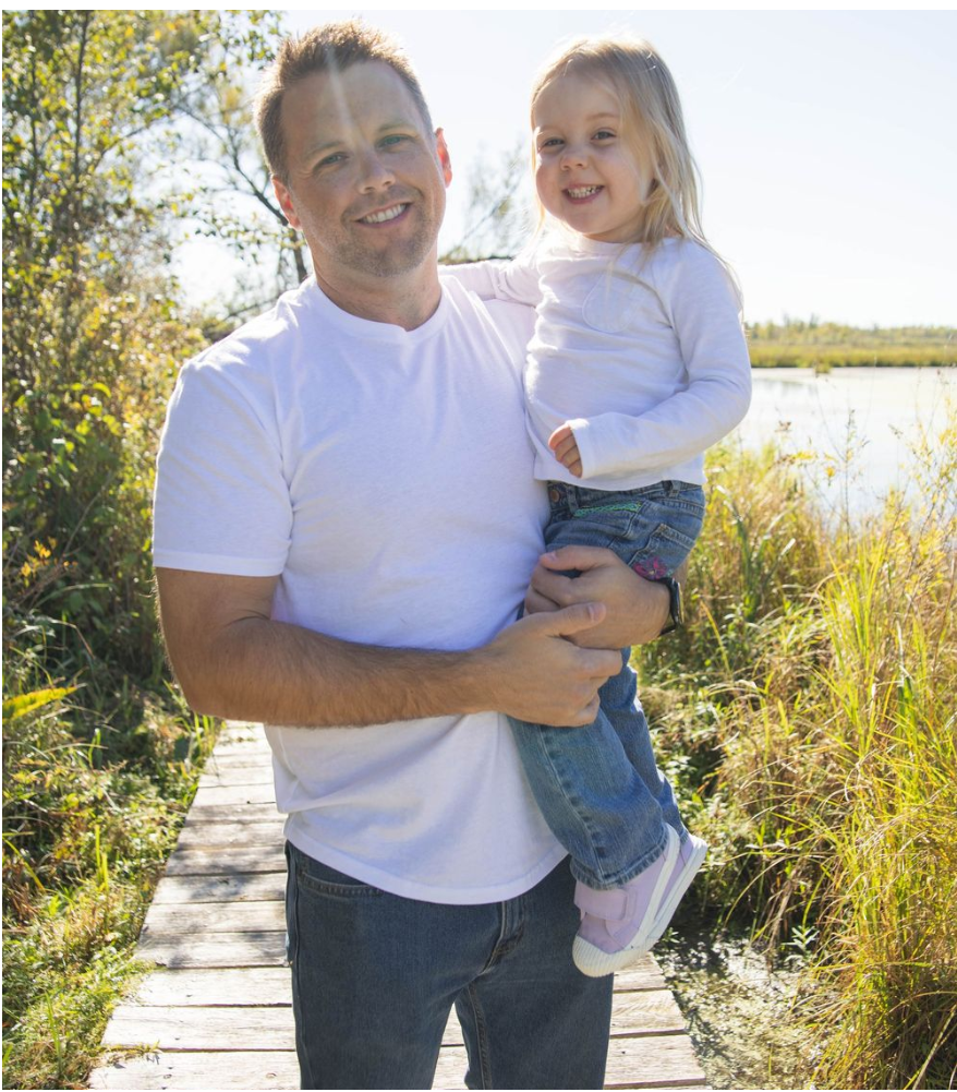 A smiling man holds a young blonde girl on a wooden boardwalk surrounded by autumn foliage near a lake.