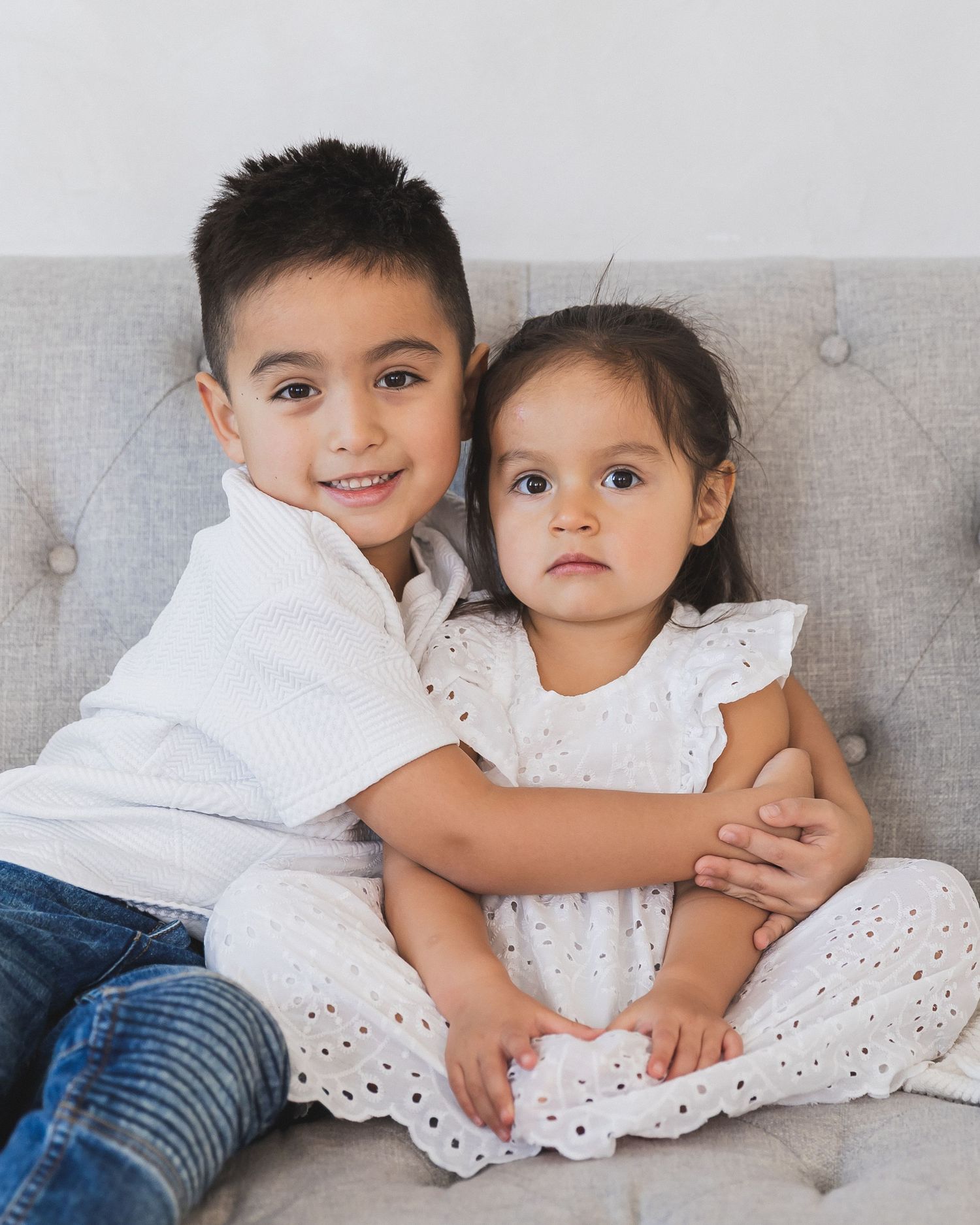 A smiling young boy lovingly hugs his little sister in a white eyelet dress while seated on a gray sofa.