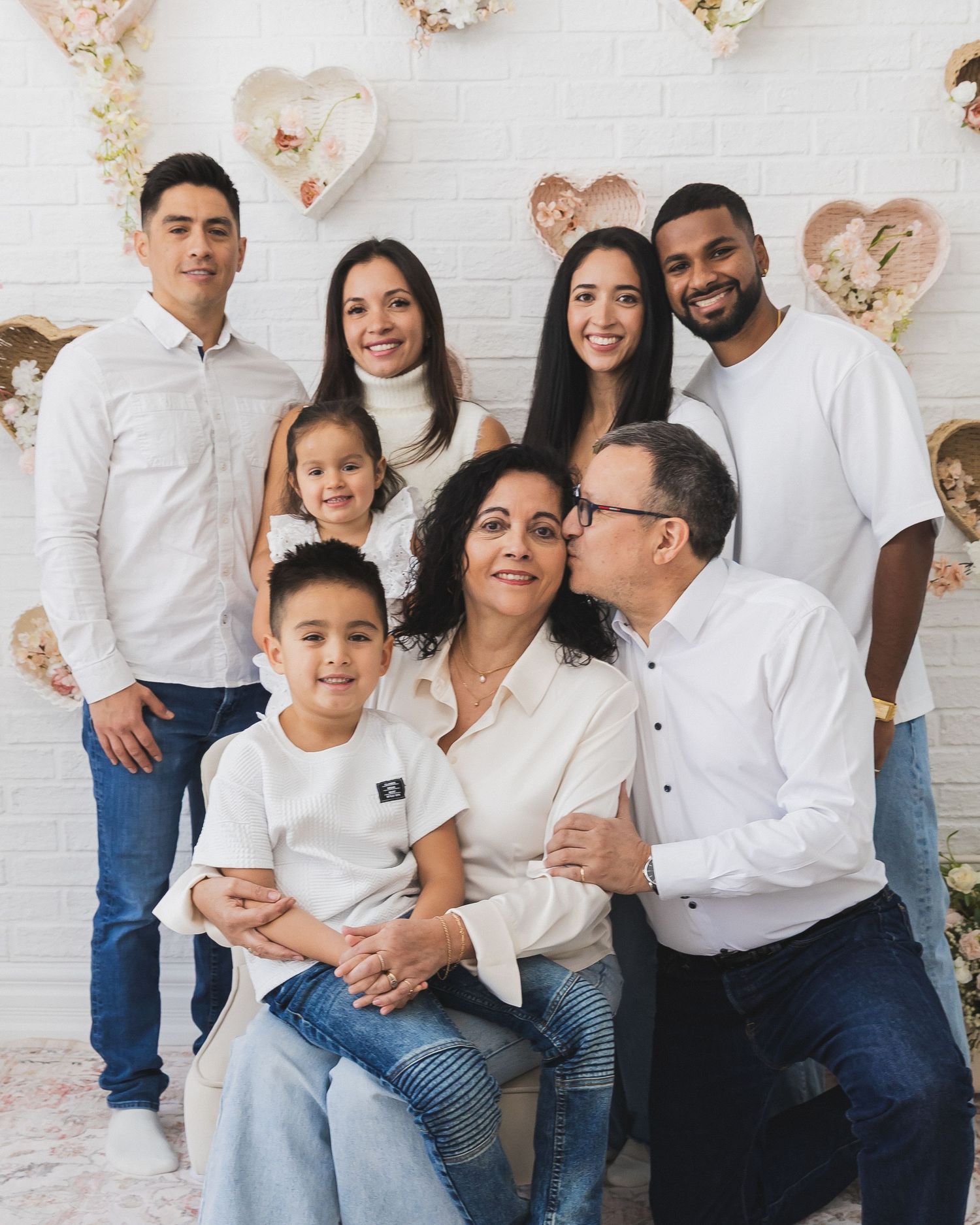 A large multigenerational Latino family gathers for a portrait with heart decorations in the background.
