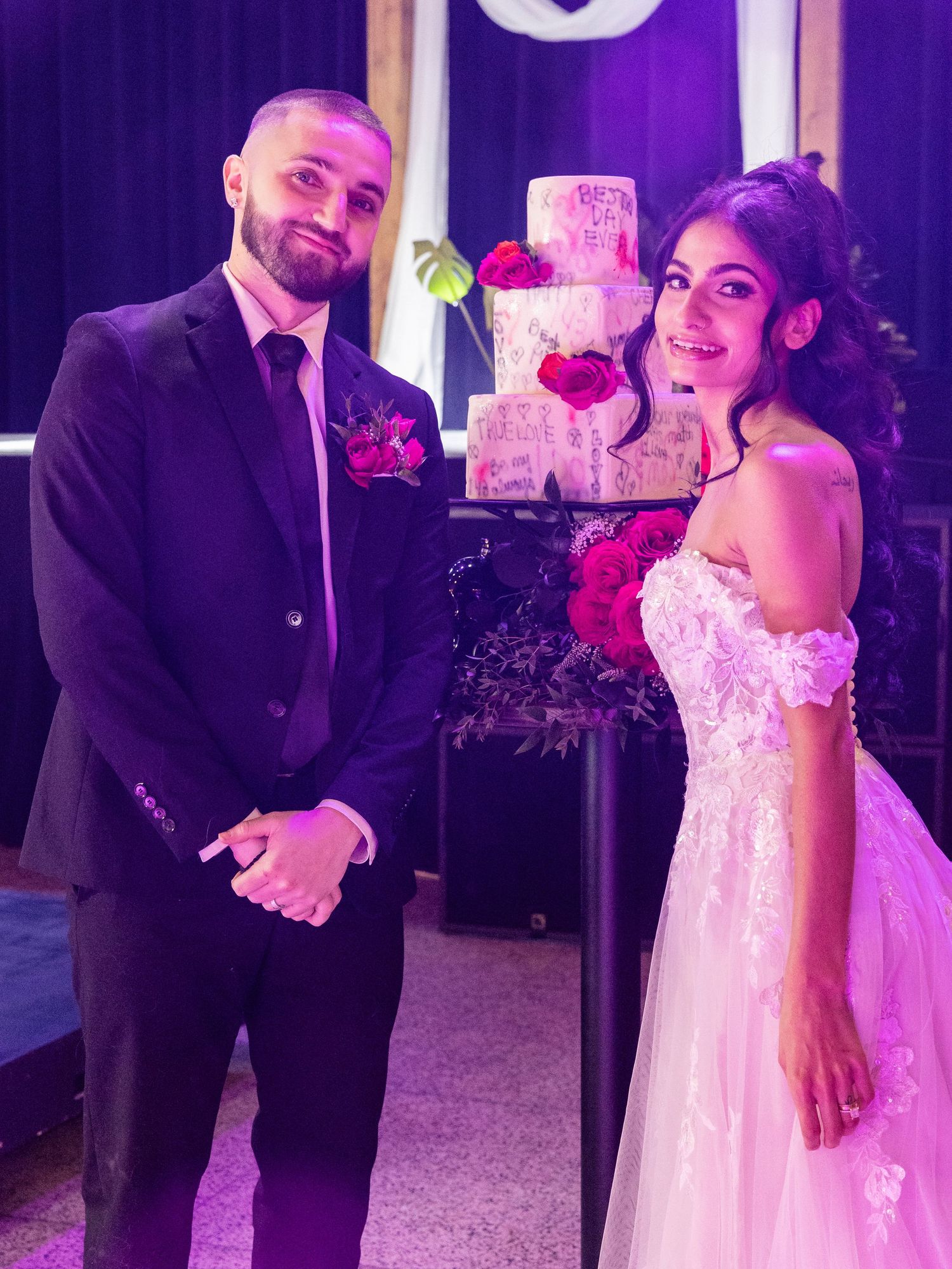 Bride and groom smile beside elegant tiered wedding cake with red roses under purple lighting.