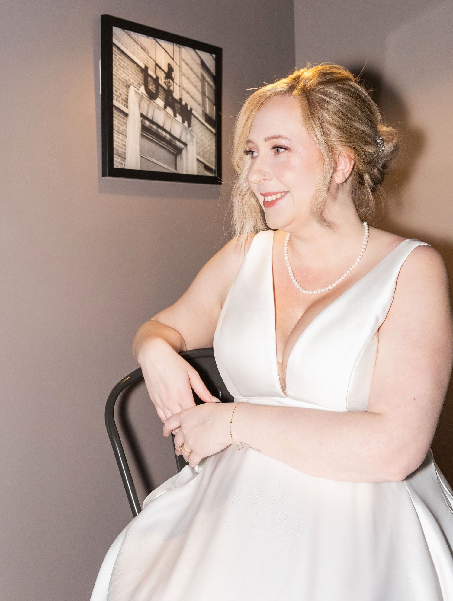 Smiling blonde bride in a white deep V-neck satin gown seated on a chair wearing a pearl necklace.
