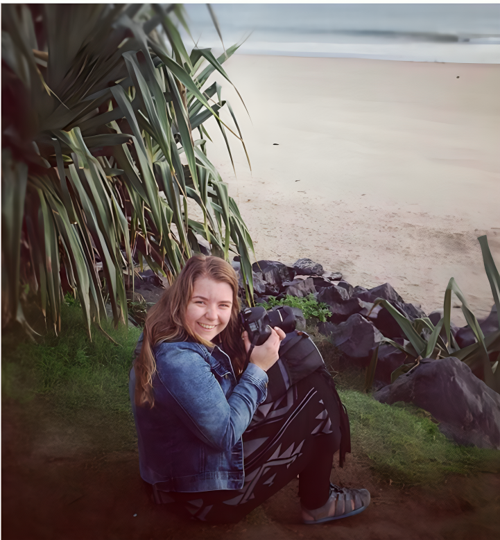 Young woman crouching with camera near tropical plants and rocks on a sandy beach shoreline.