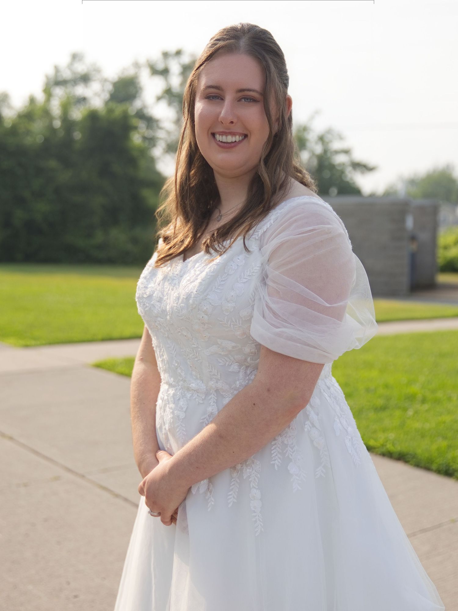 Smiling bride in white lace ball gown with sheer puff sleeves poses outdoors on a sunny day.