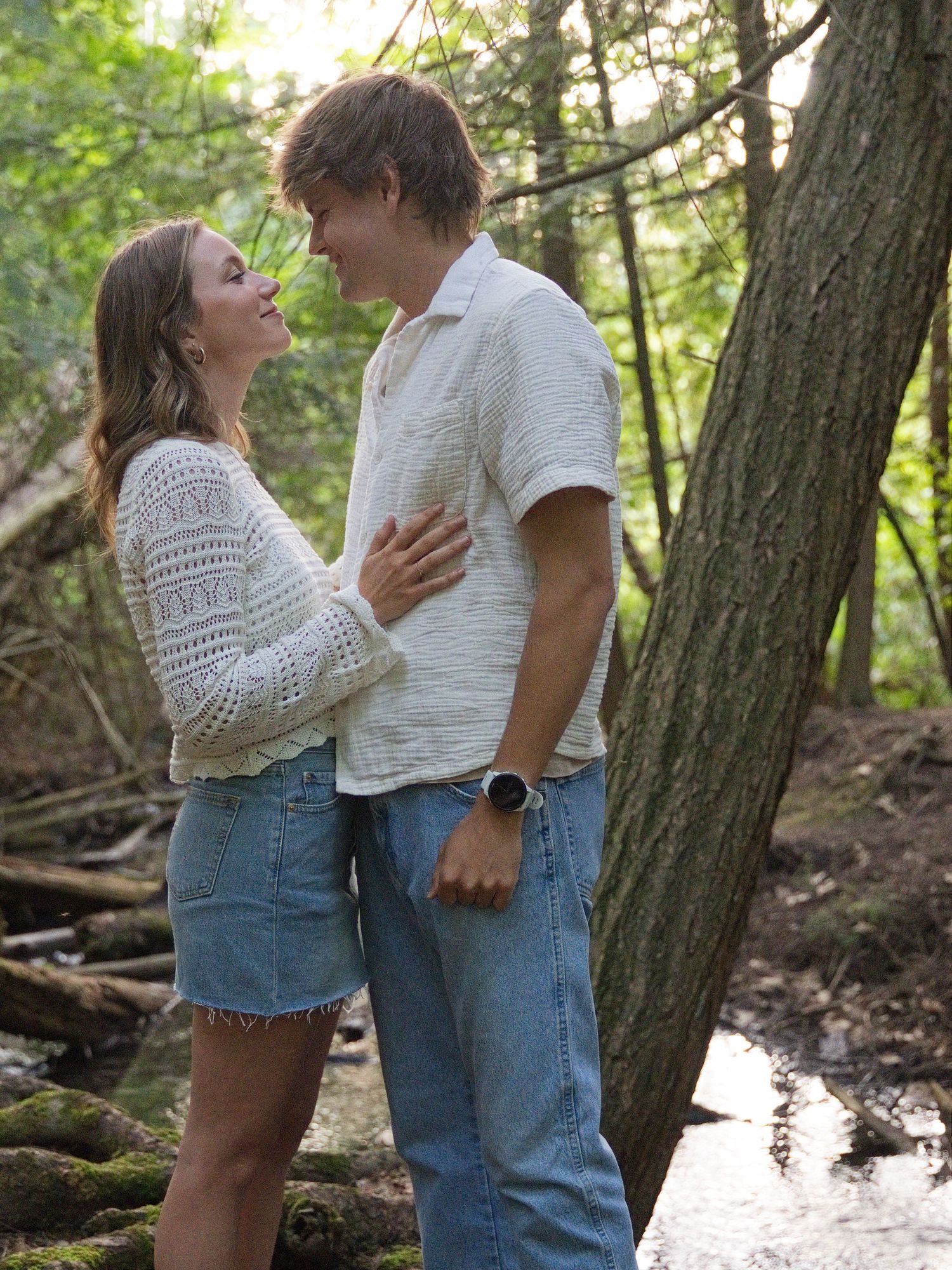 A young couple shares a romantic moment in a lush forest setting, dressed in casual white tops and denim.
