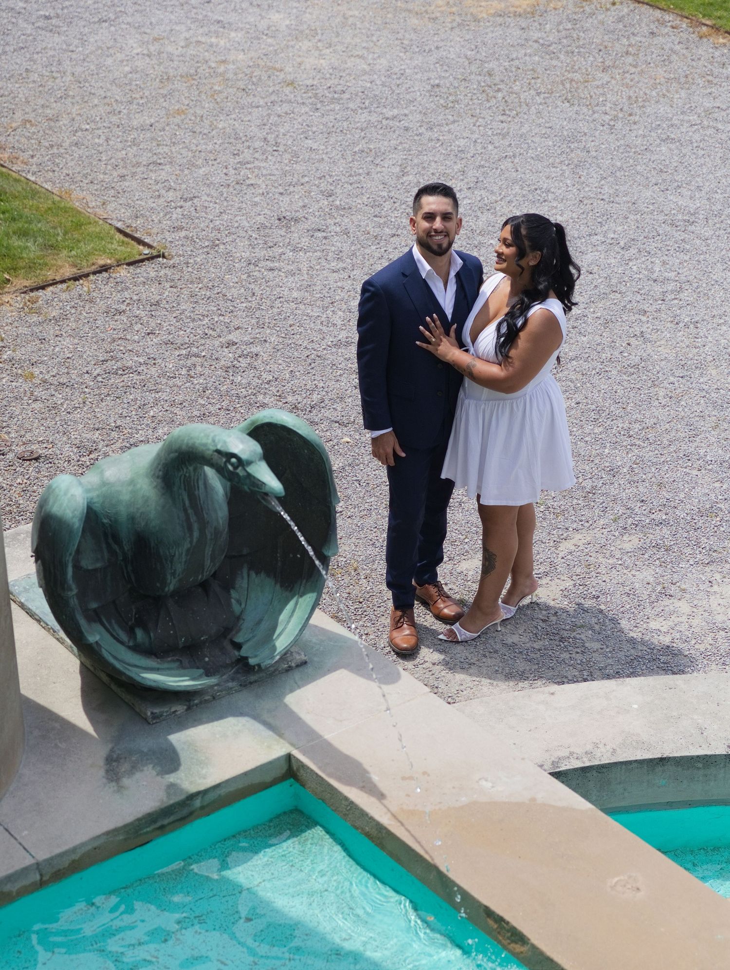 Couple posing romantically near a bronze fountain sculpture by a turquoise pool in an elegant outdoor setting.