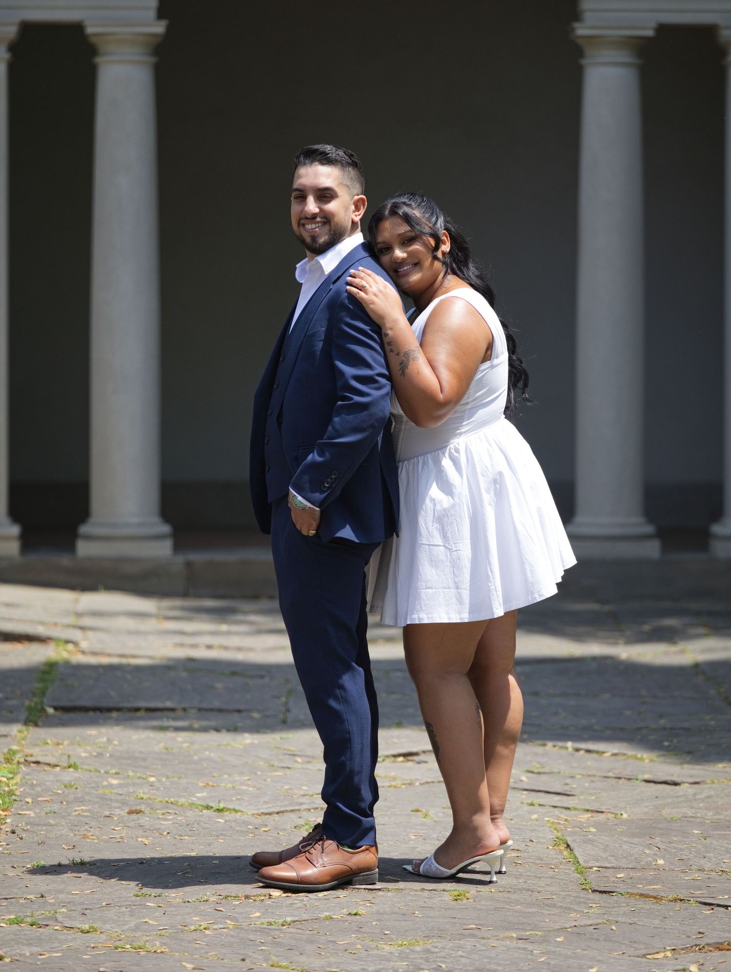 Couple posing back-to-back outdoors, man in navy suit, woman in white dress, near classical columns.