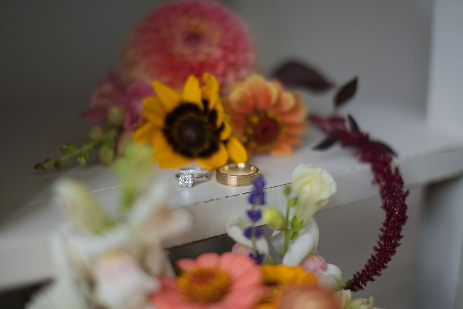 Wedding rings rest among colorful bouquet featuring sunflowers, dahlias, and wildflowers in pink, yellow, and orange hues.