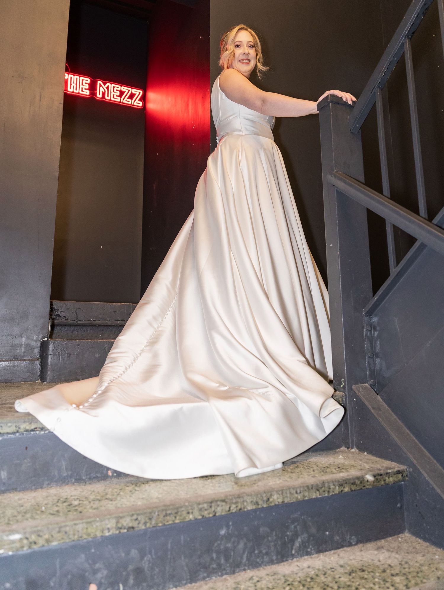 Bride twirls in a stunning champagne satin ball gown on a staircase near a neon sign.