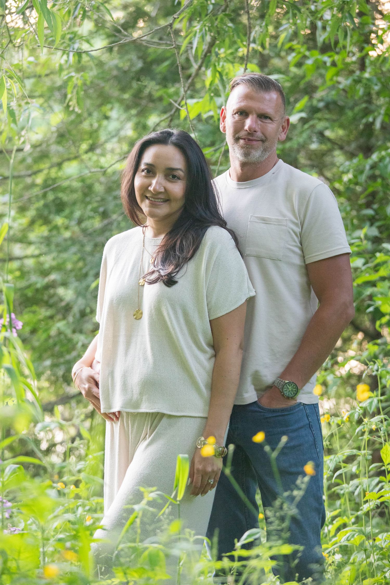 A smiling couple stands together outdoors among lush green foliage, both wearing light-colored casual clothing.