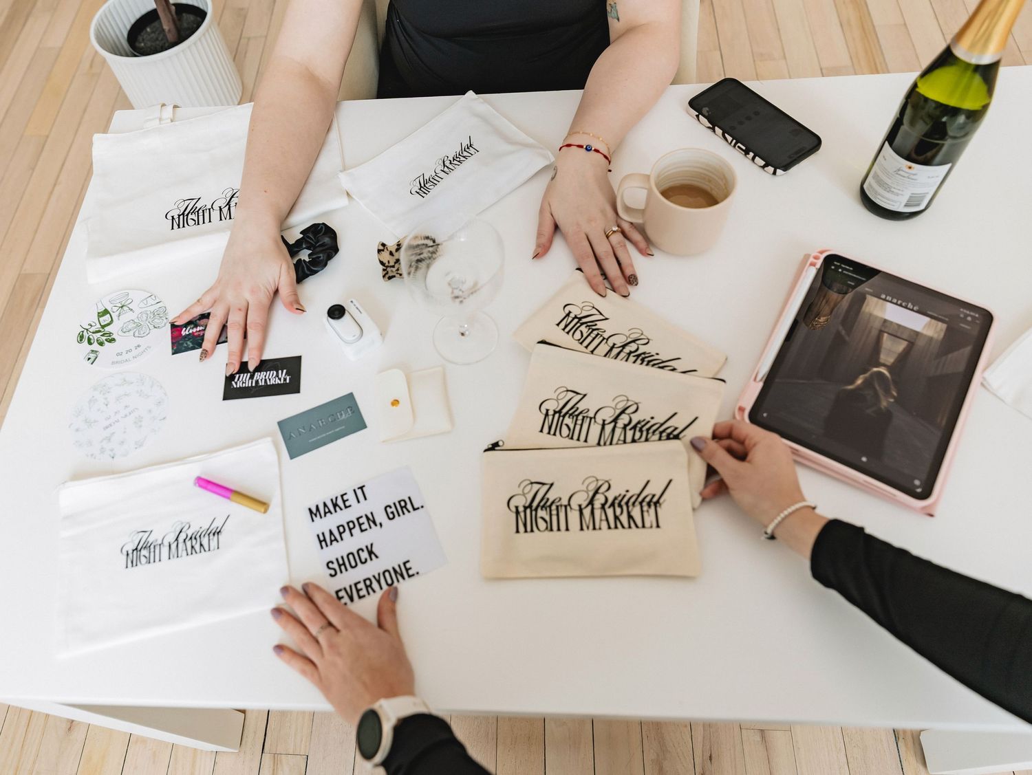 Two women collaborate at a white desk with calligraphy pouches, tablets, and champagne in a creative workspace.