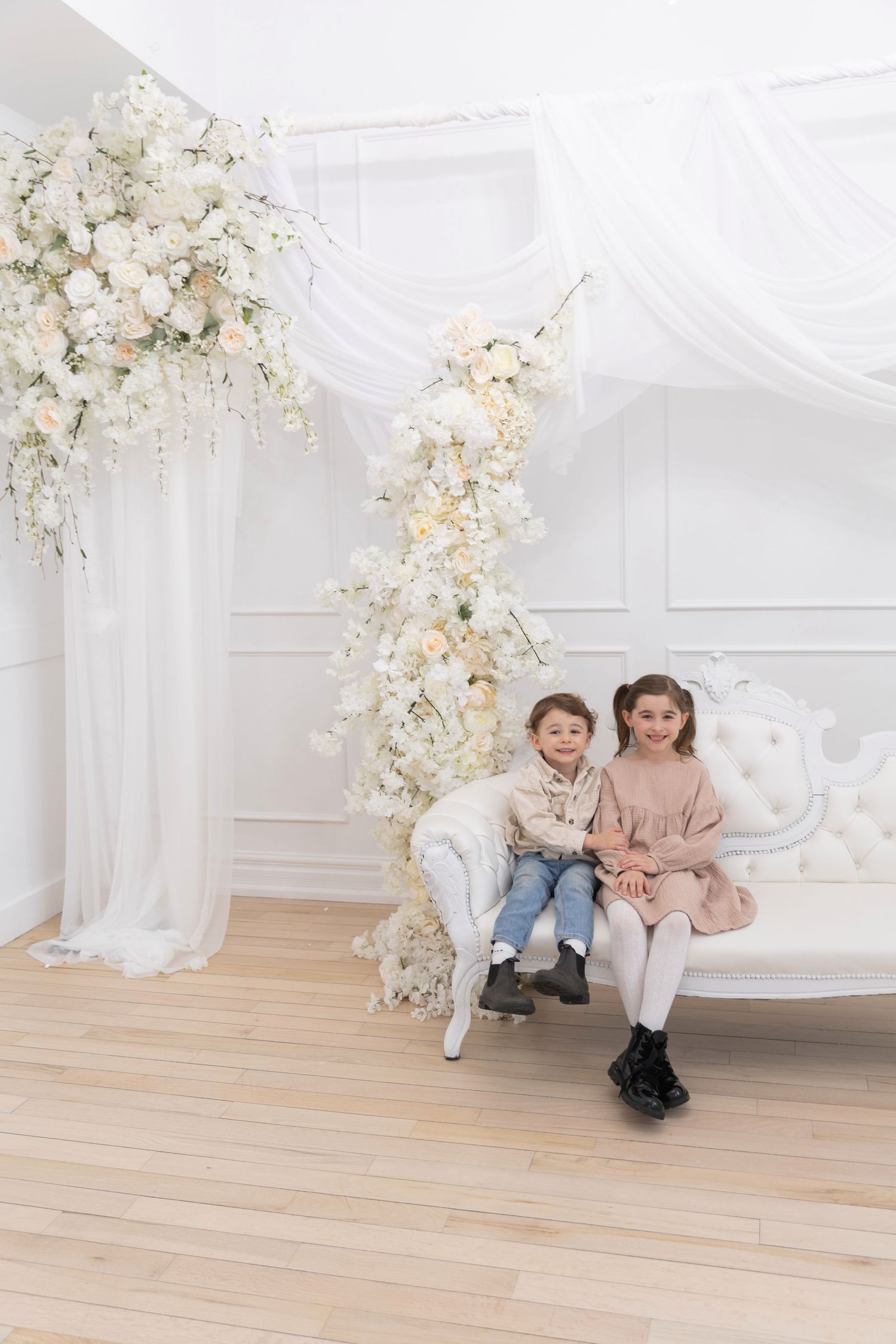 Two children sitting on white ornate bench surrounded by elegant white floral decorations.