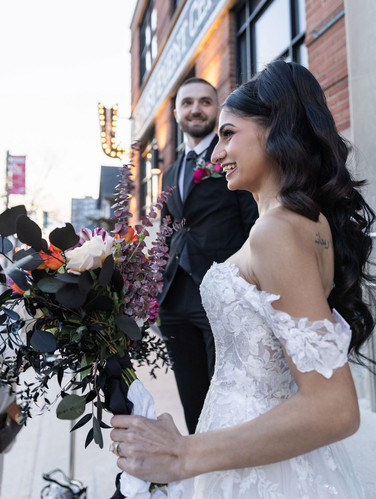 Bride holding lush floral bouquet with groom standing behind her on urban brick street setting.