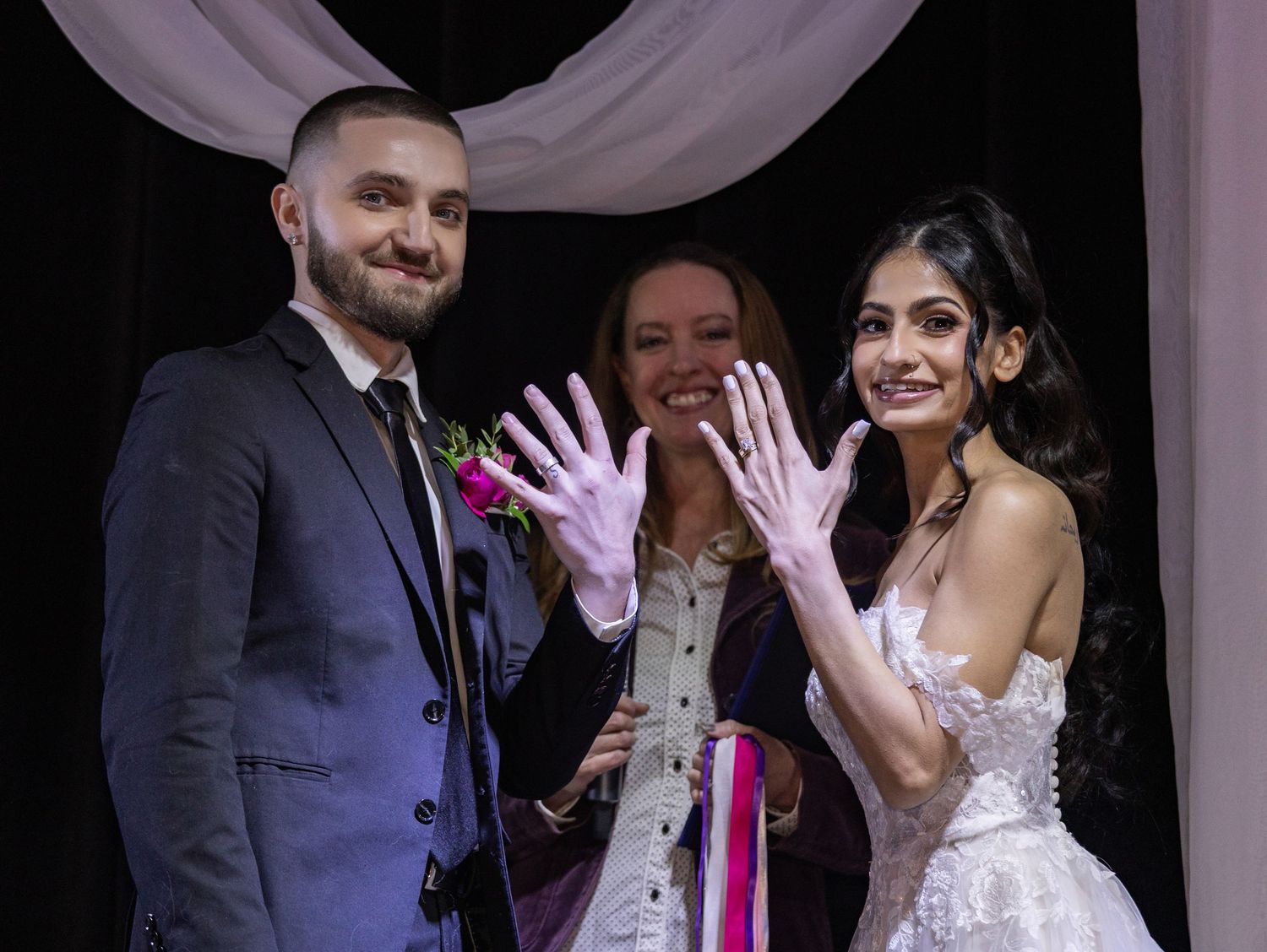Bride and groom showing pink ribbon hand-fasting on hands with officiant at wedding ceremony.