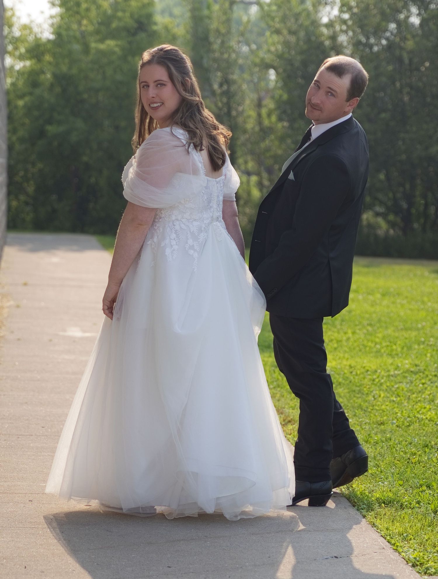Bride and groom in wedding attire pose back-to-back on a sunlit outdoor pathway.