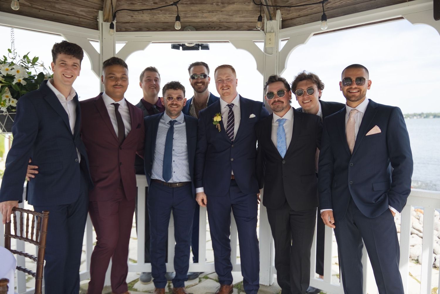 Group of nine men in suits posing together under a gazebo at a waterfront wedding celebration.