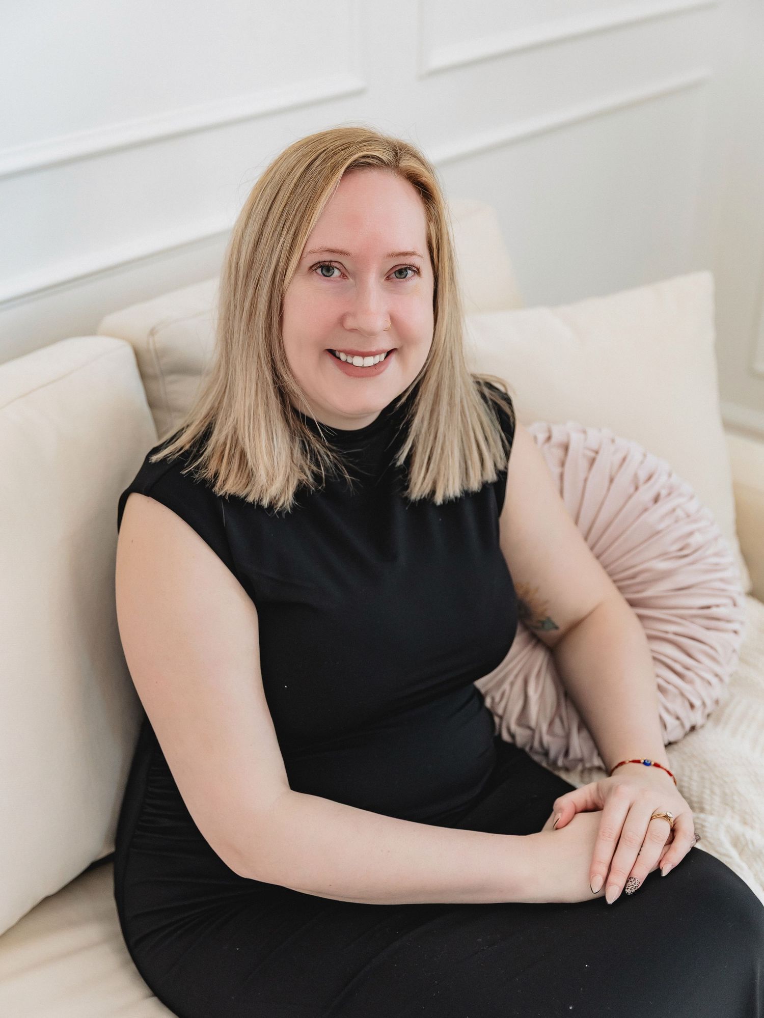 Smiling blonde woman in black sleeveless turtleneck sitting on a cream sofa with a pink pillow.