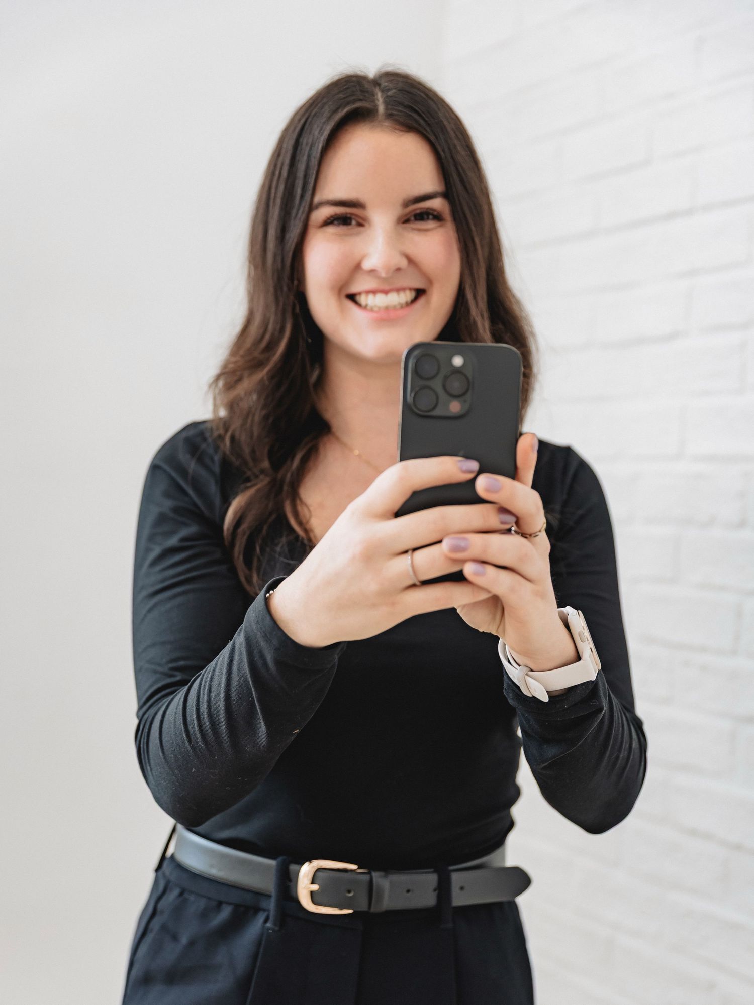 Smiling brunette woman in black outfit taking a selfie with iPhone against white brick wall.