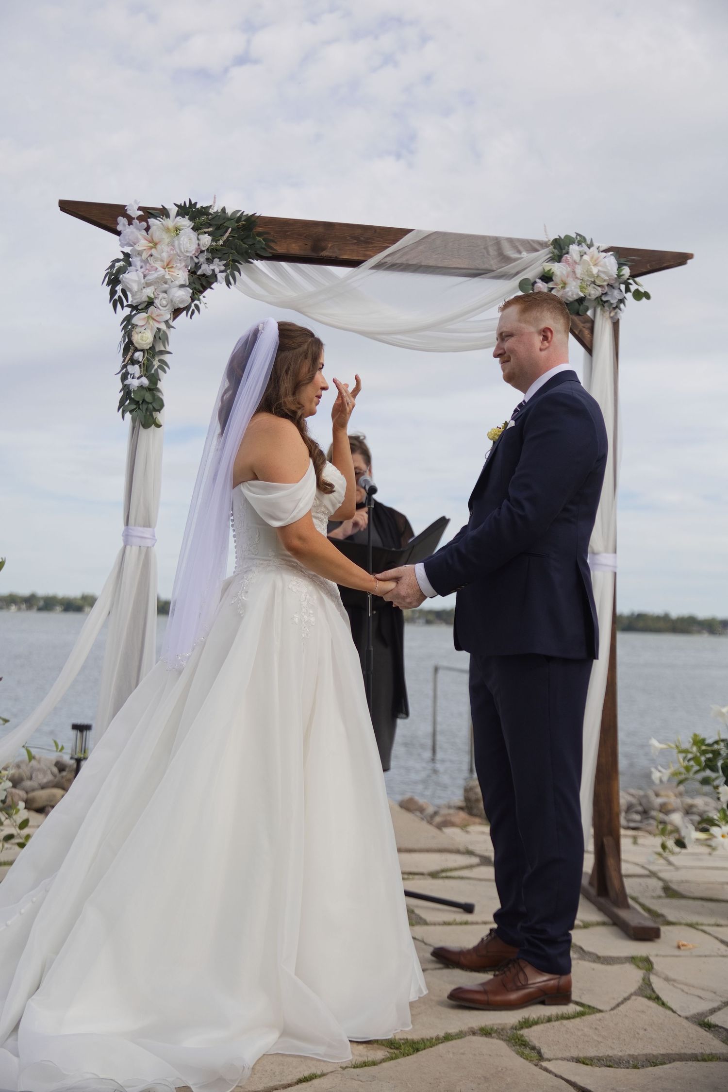 Bride and groom exchange vows under a floral arch by the waterfront during an outdoor wedding ceremony.