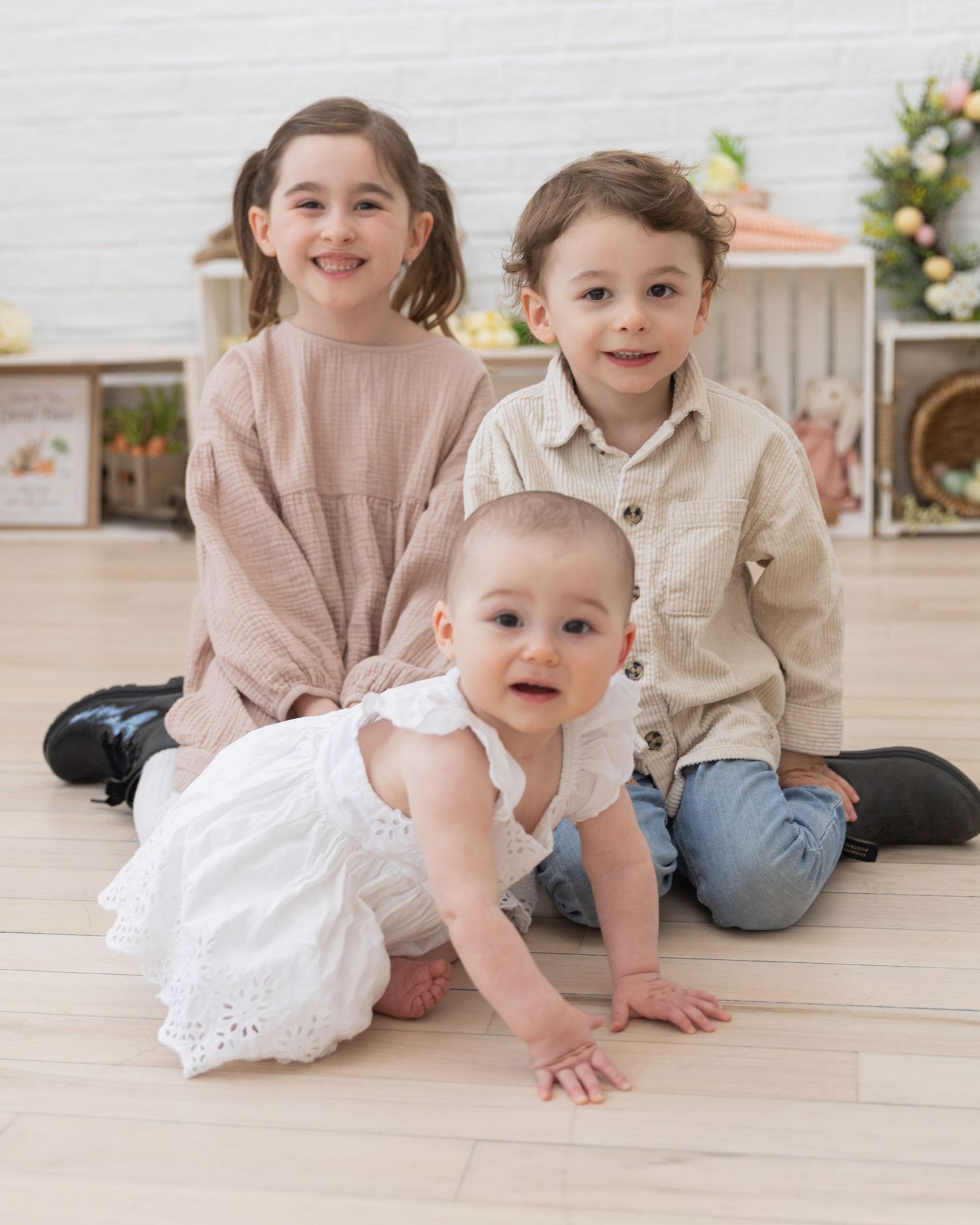 Three young siblings on floor with Easter backdrop, baby crawling in white eyelet dress.