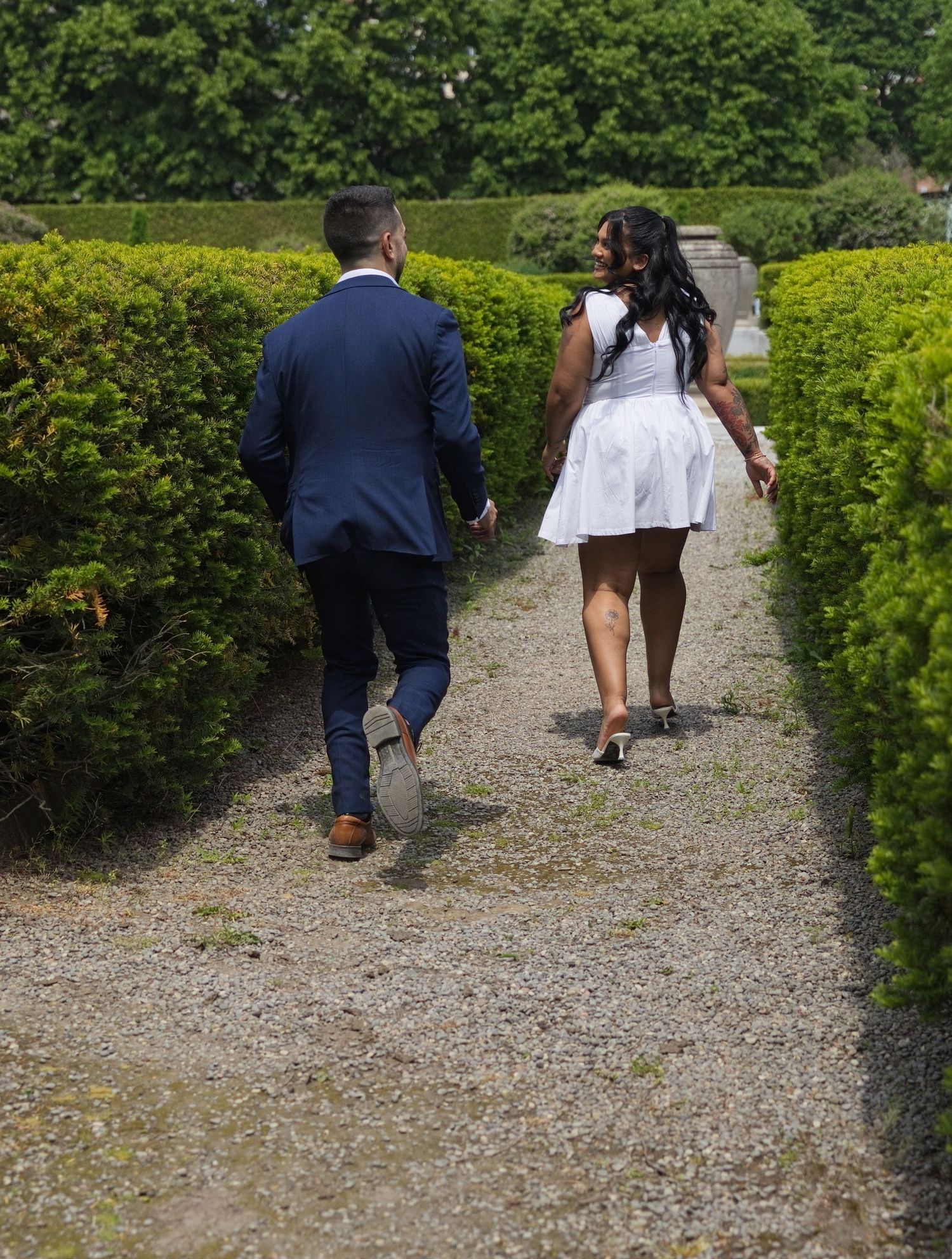 Couple in wedding attire walking hand in hand through a lush green hedge garden pathway on a sunny day.