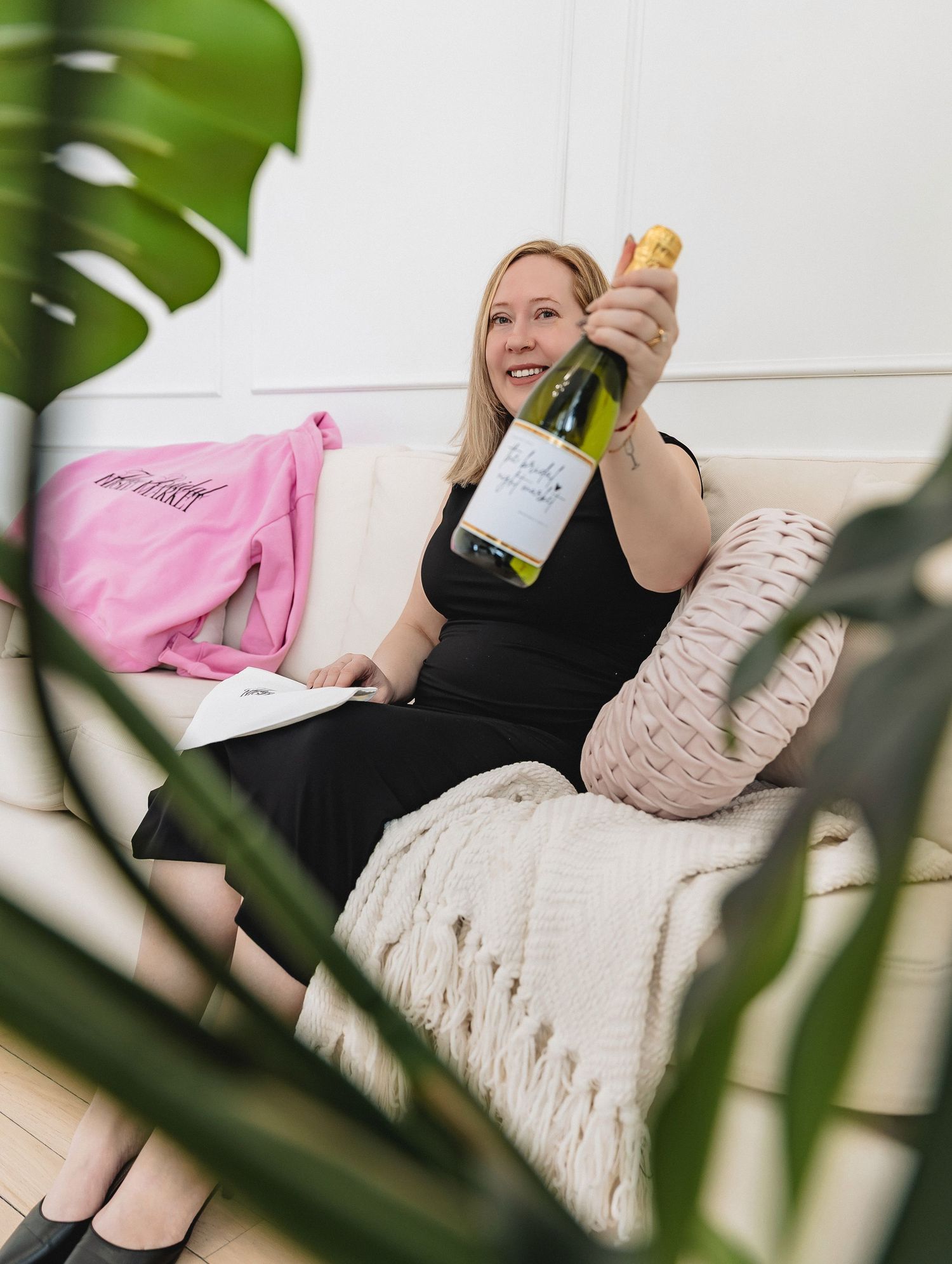 Smiling blonde woman in black dress holding wine bottle while relaxing on white sofa with pink bag and plants nearby.