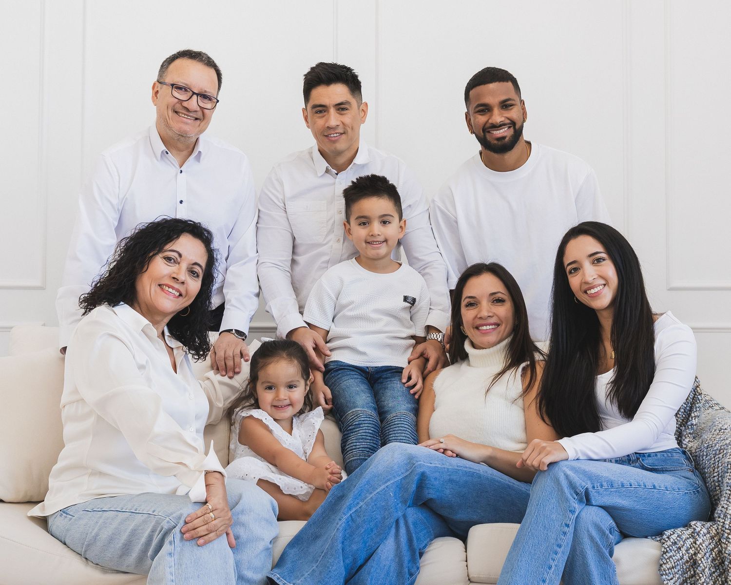 Large multigenerational family smiling together on a white couch in a bright studio.