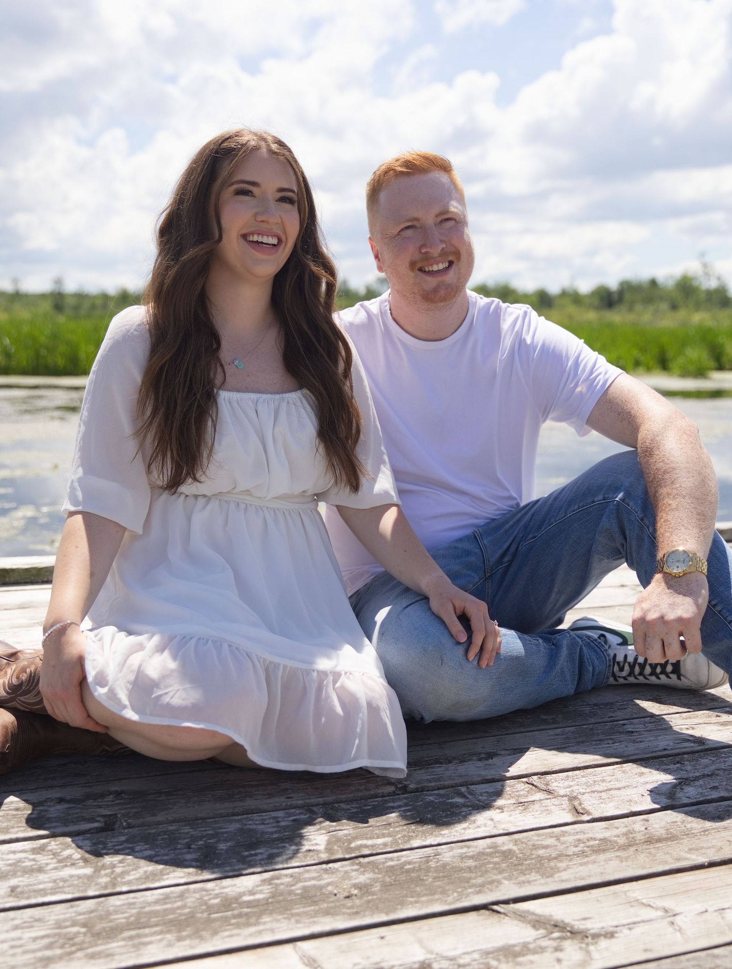 Happy couple sitting on a wooden dock by a lake, woman in white dress and man in white shirt and jeans.