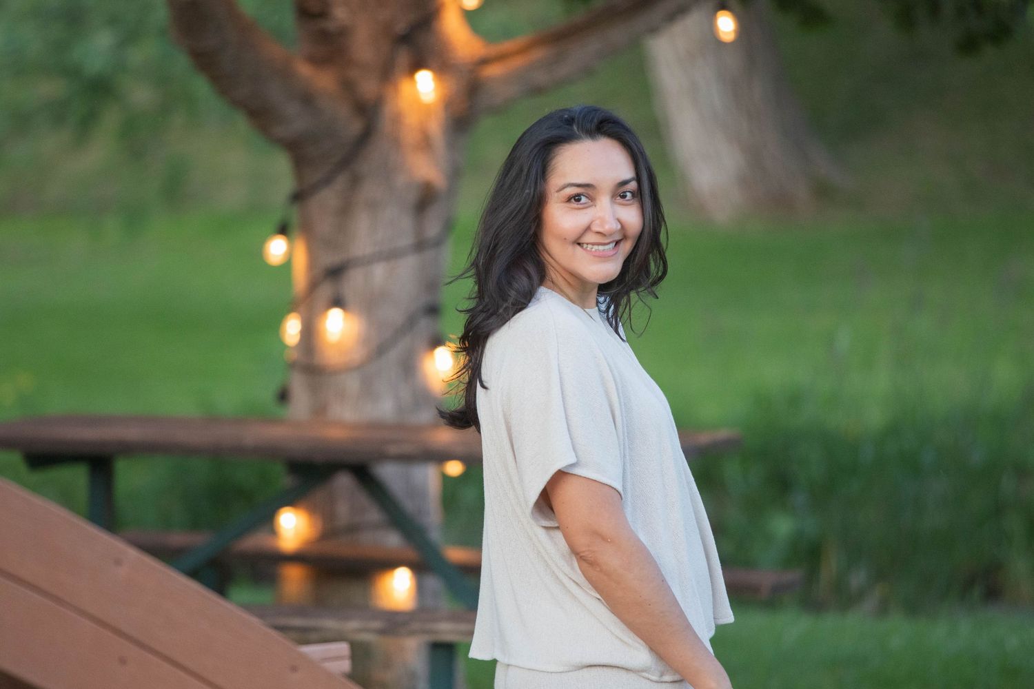 Smiling woman in beige top posing outdoors near string lights and a tree at dusk.
