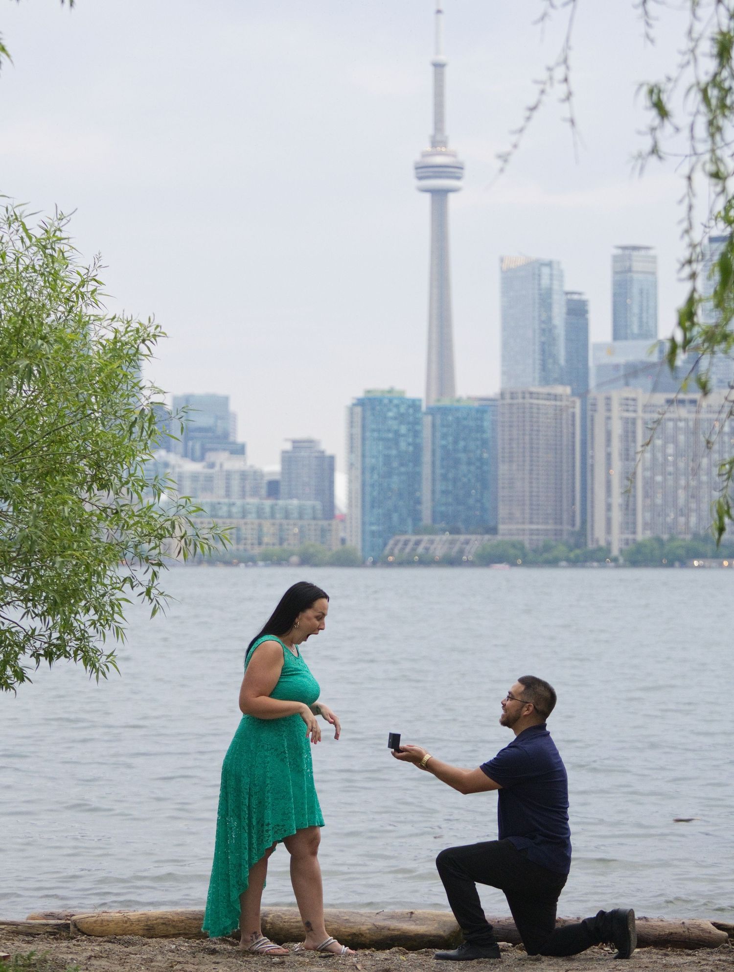 Man proposes on one knee to woman in green dress by Toronto waterfront with CN Tower skyline backdrop.