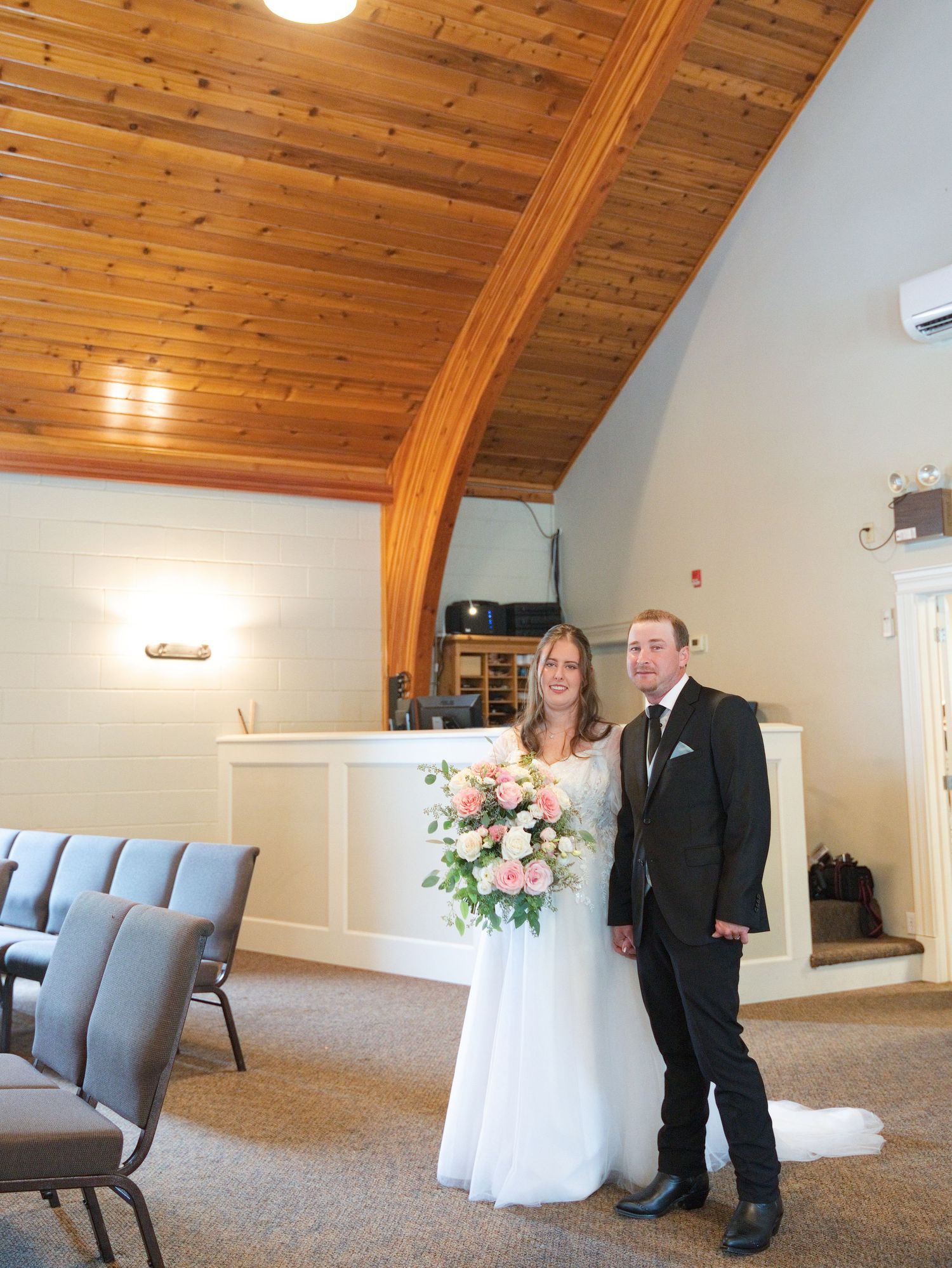 Bride in white gown holding pink bouquet stands with groom in black suit inside a wooden ceiling chapel.