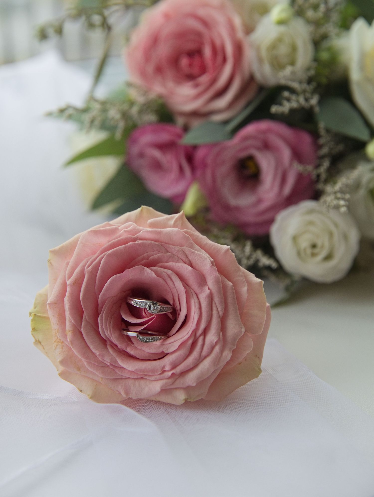 A diamond engagement ring nestled inside a pink rose bloom with a soft bouquet of pink and white flowers in the background.