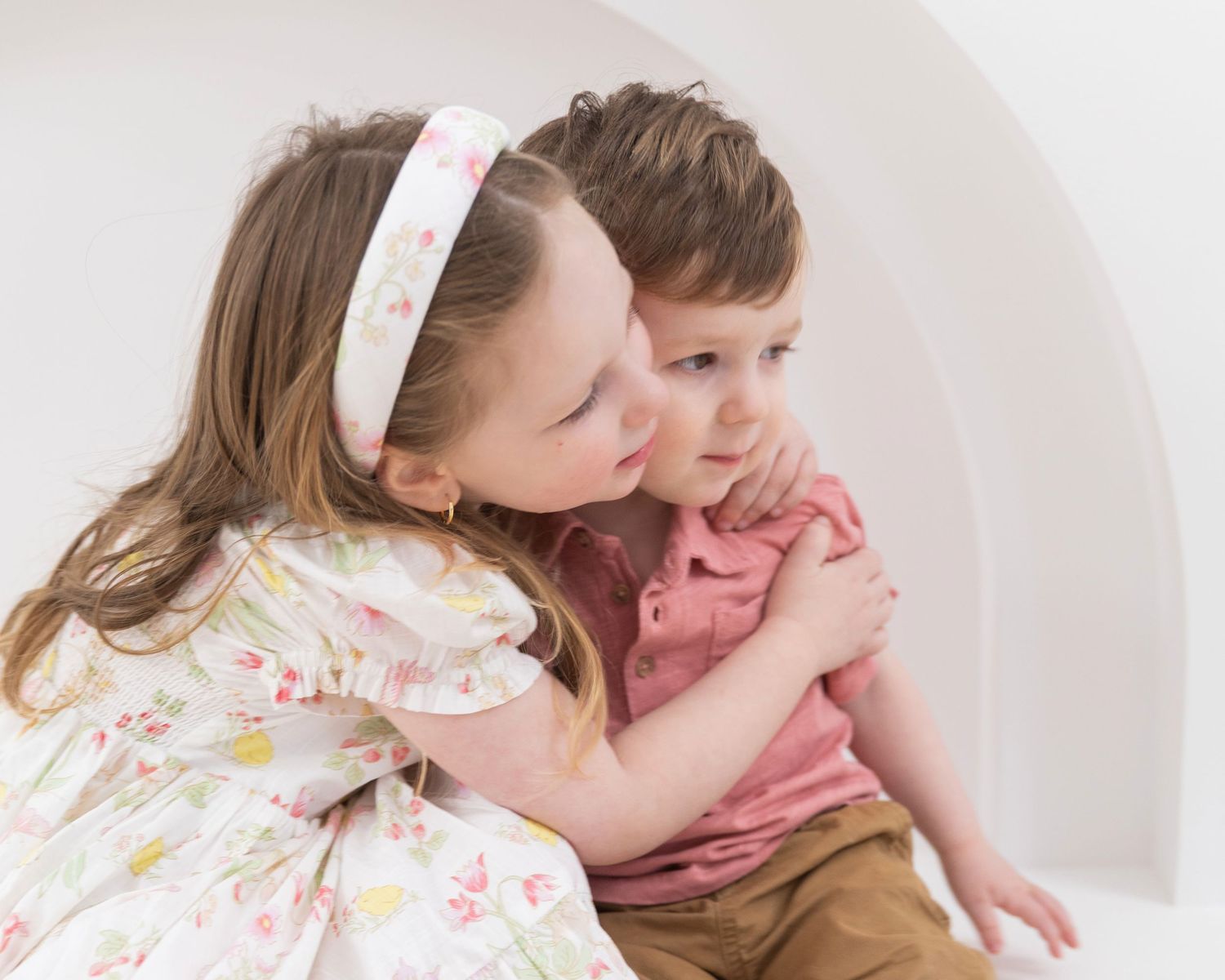 Sweet older sister giving her younger brother a gentle kiss on the cheek in a bright white setting.