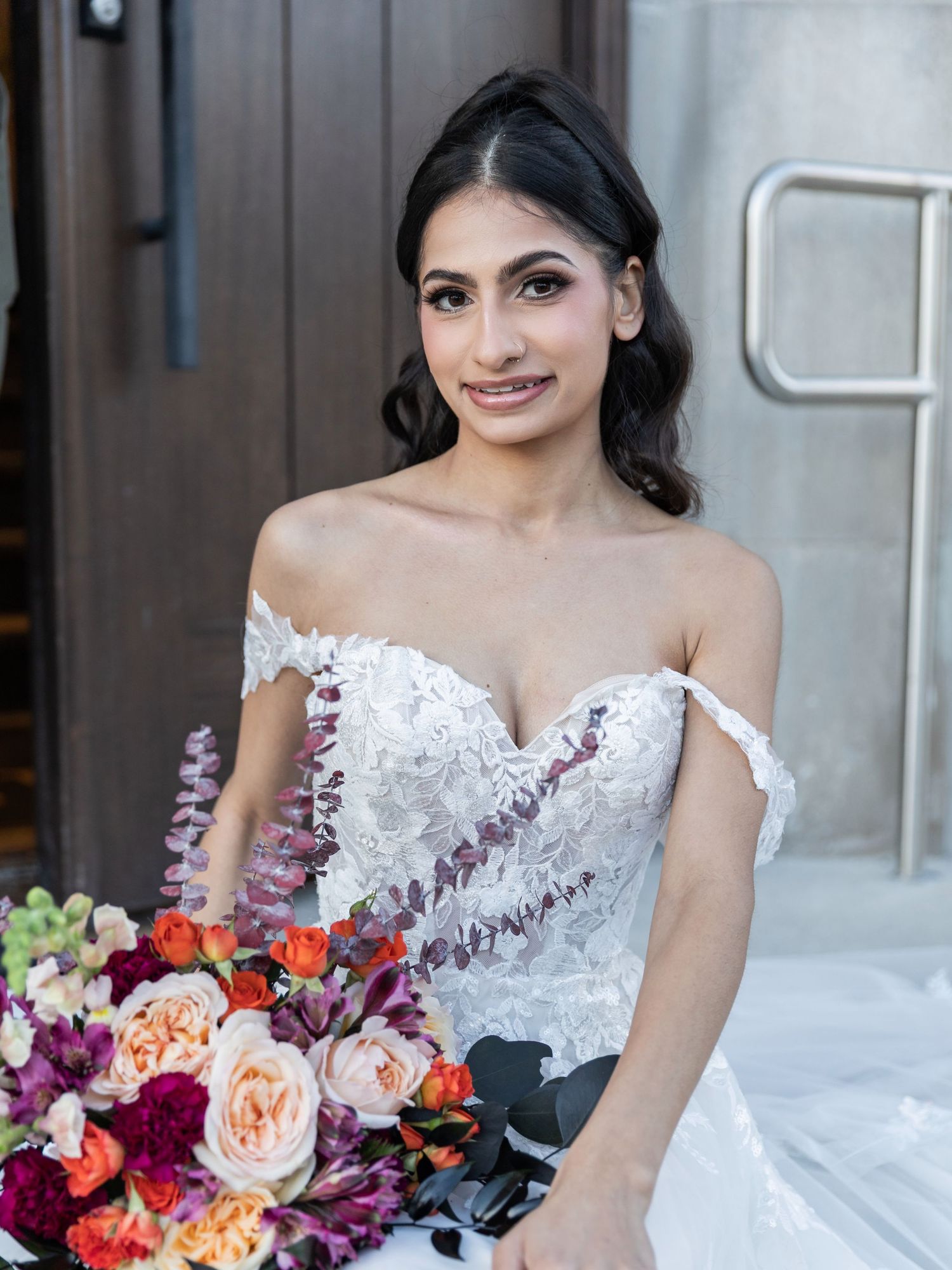 Beautiful bride in off-shoulder lace wedding gown holding vibrant colorful bridal bouquet outdoors.