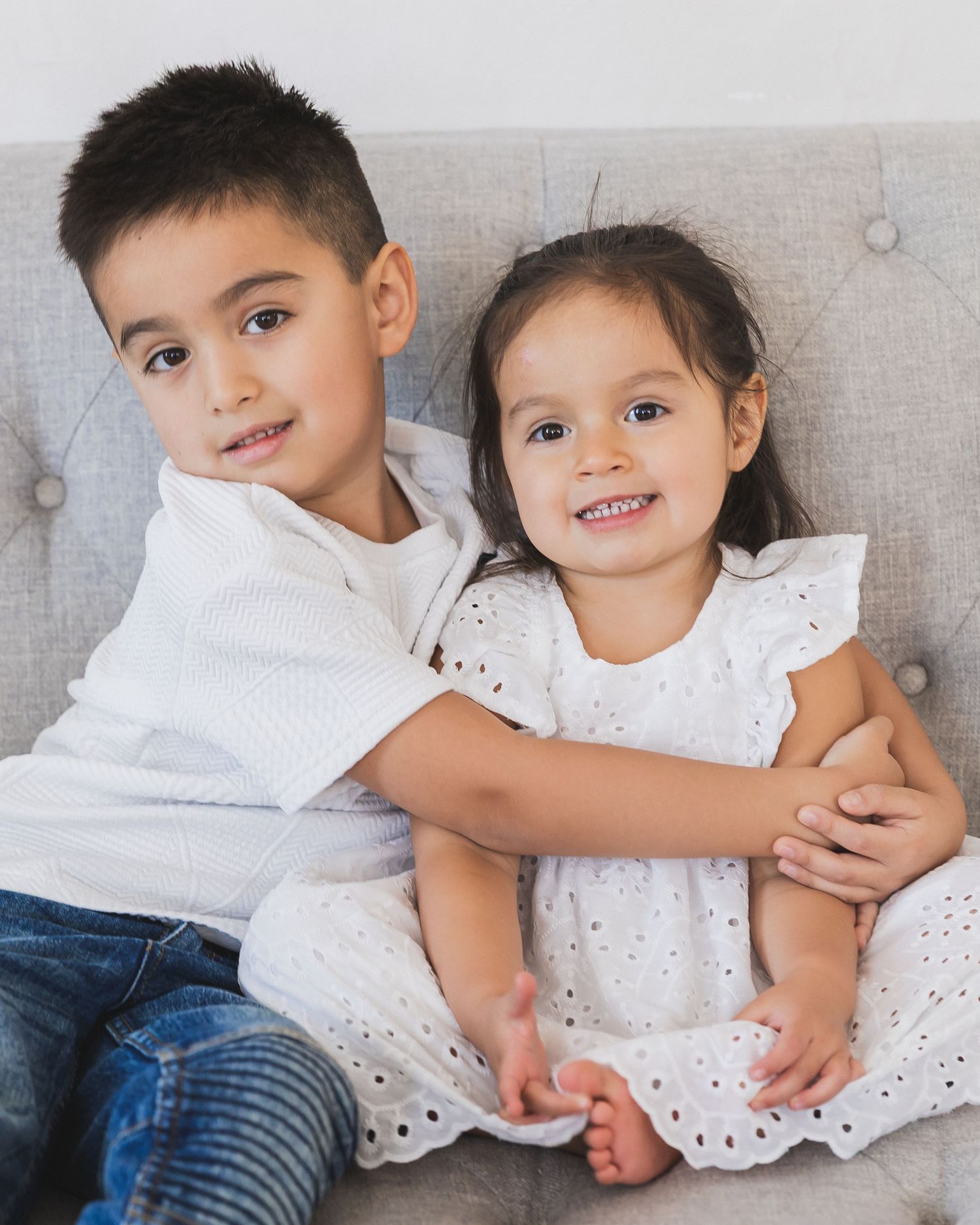 Young boy hugging his smiling little sister in white eyelet dress on gray couch.