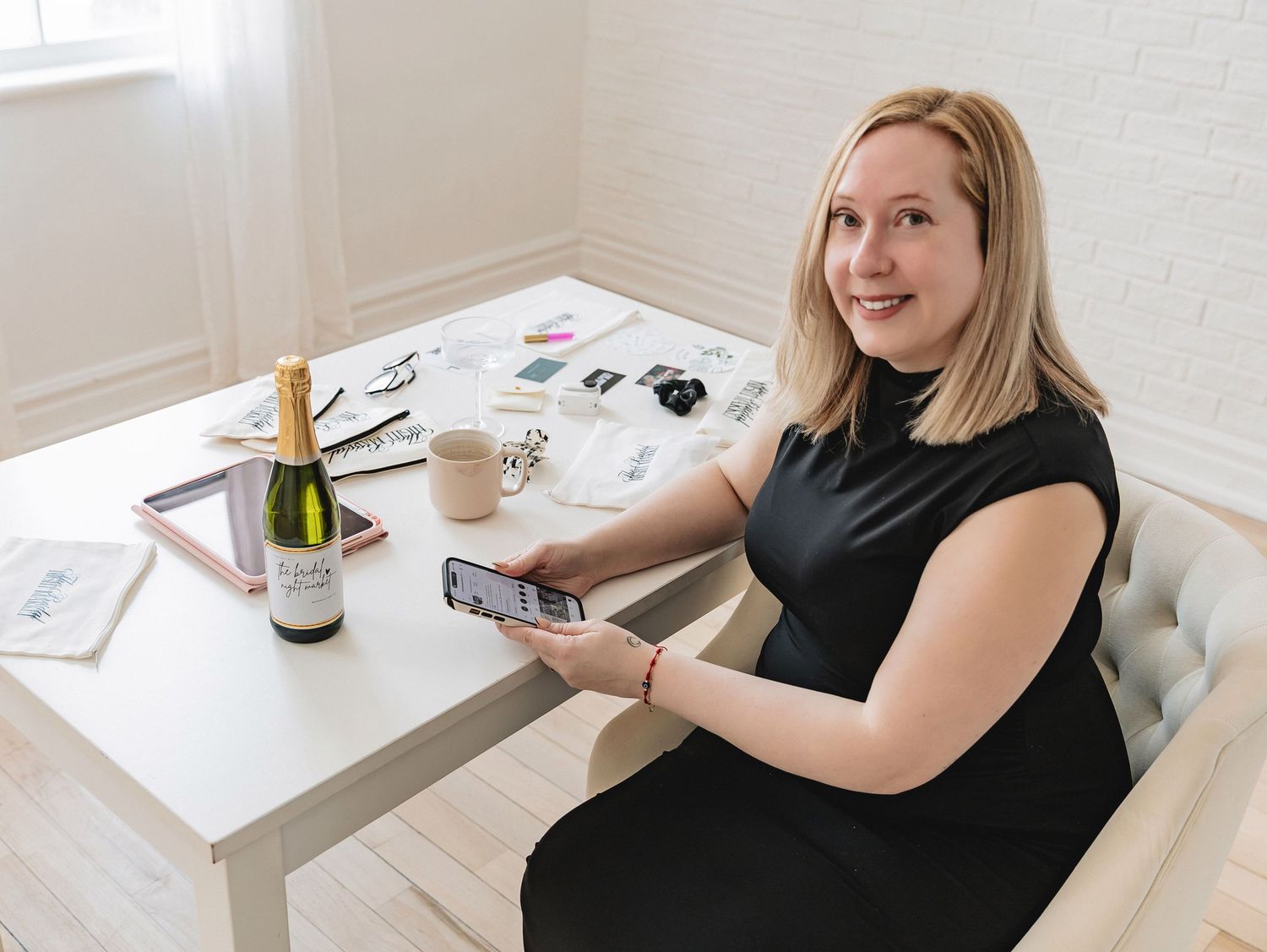Woman in black dress sitting at white desk with champagne bottle, coffee, and phone in bright office.