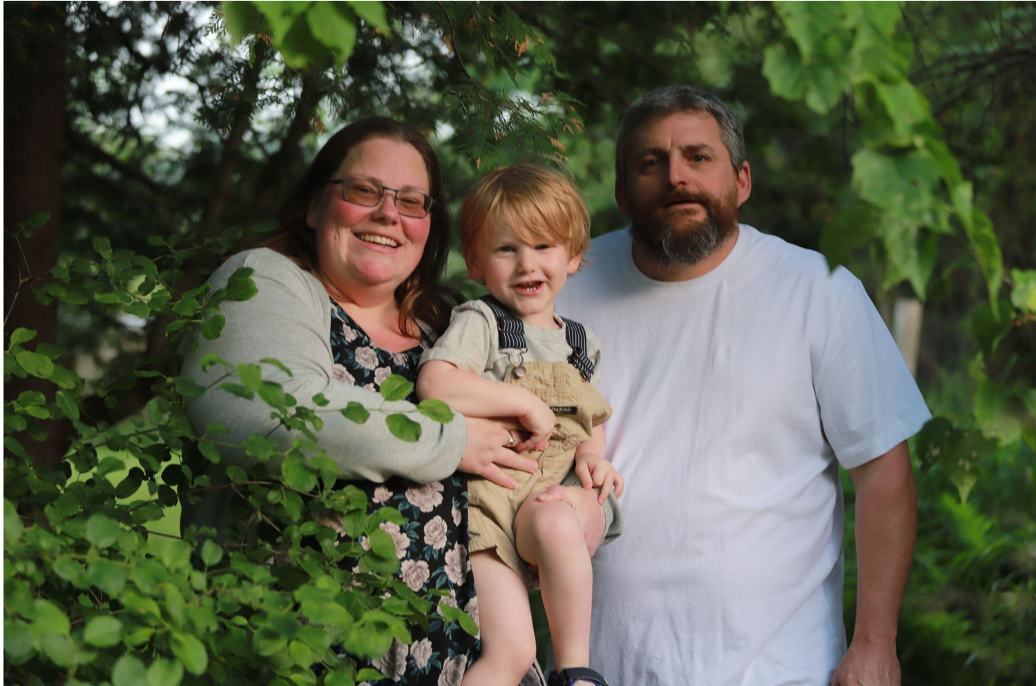 A smiling family of three poses outdoors, with parents holding a young toddler surrounded by lush green trees.