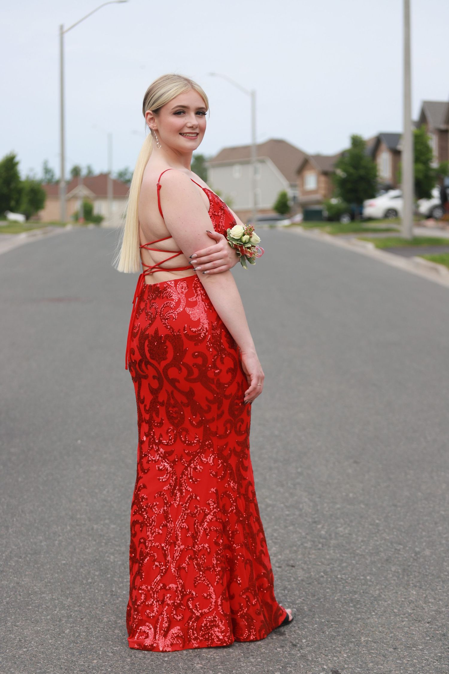 Young woman in a red sequin prom gown with floral pattern poses on suburban street wearing a wrist corsage.