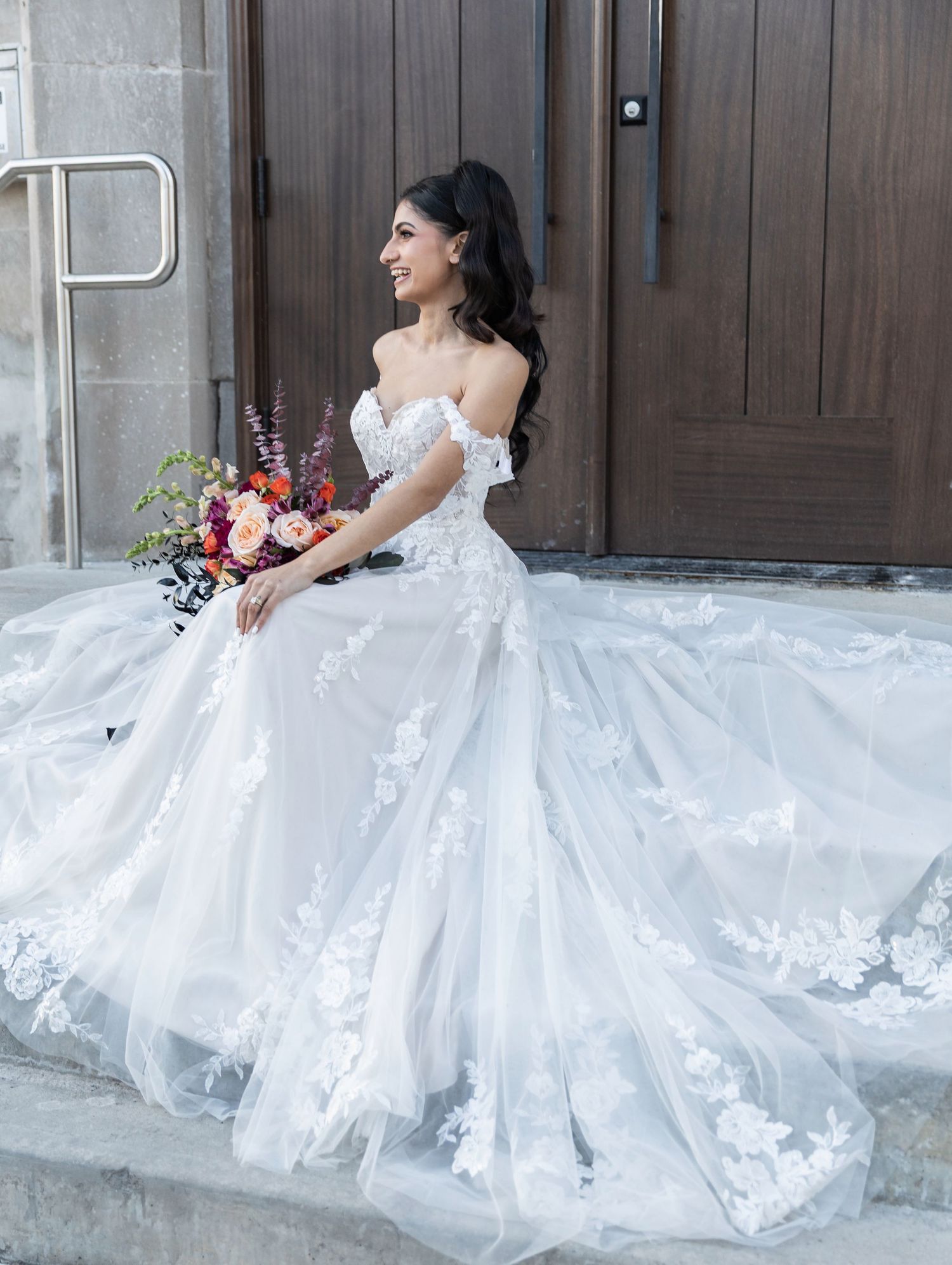Bride in off-shoulder lace ball gown sits on steps holding a colorful floral bouquet.