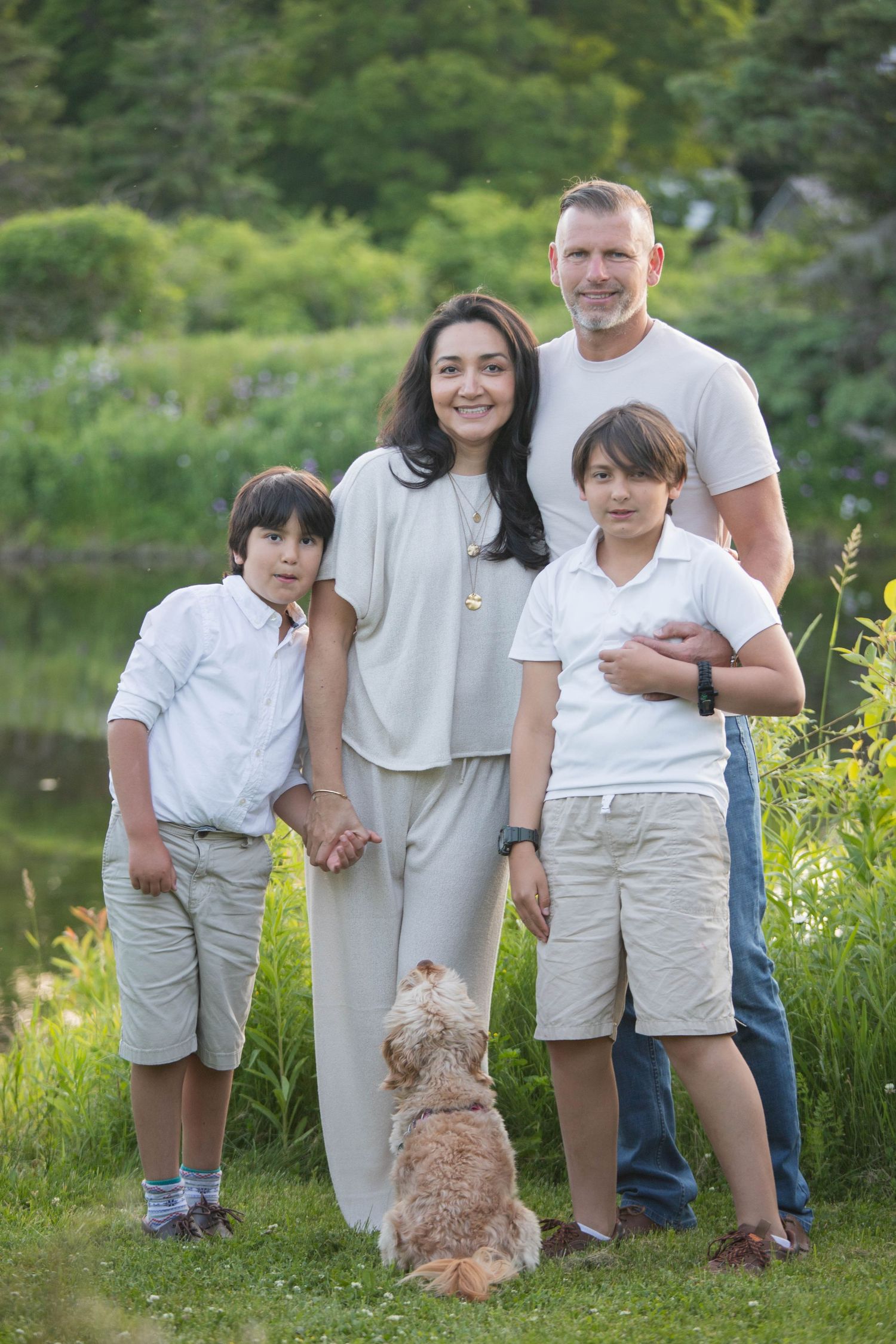 A family of four with two boys and a fluffy dog poses outdoors near a pond surrounded by greenery.