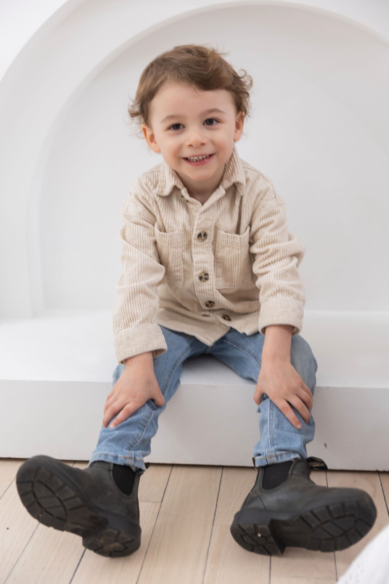 Smiling toddler boy in beige corduroy shirt, jeans, and black boots sitting on white bench.