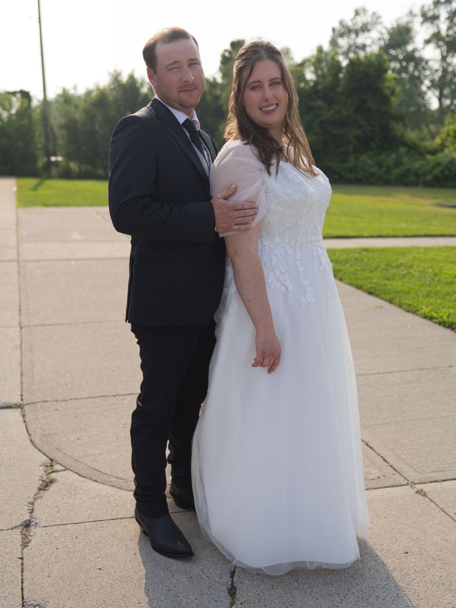 Bride in white gown and groom in black suit posing together outdoors on a sunny day at their wedding.
