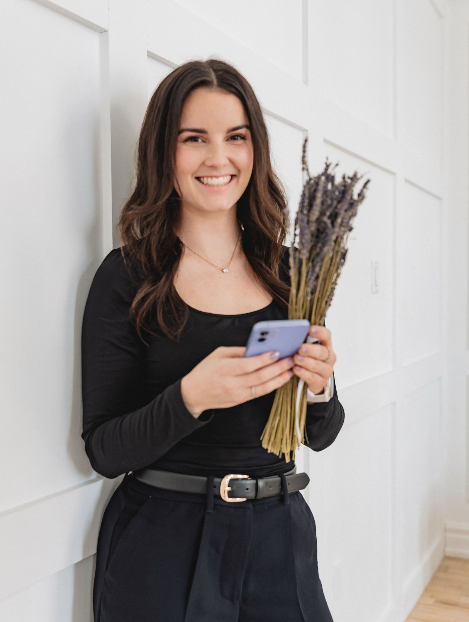 Young woman in black outfit holding lavender bouquet and smartphone, smiling against white wall.