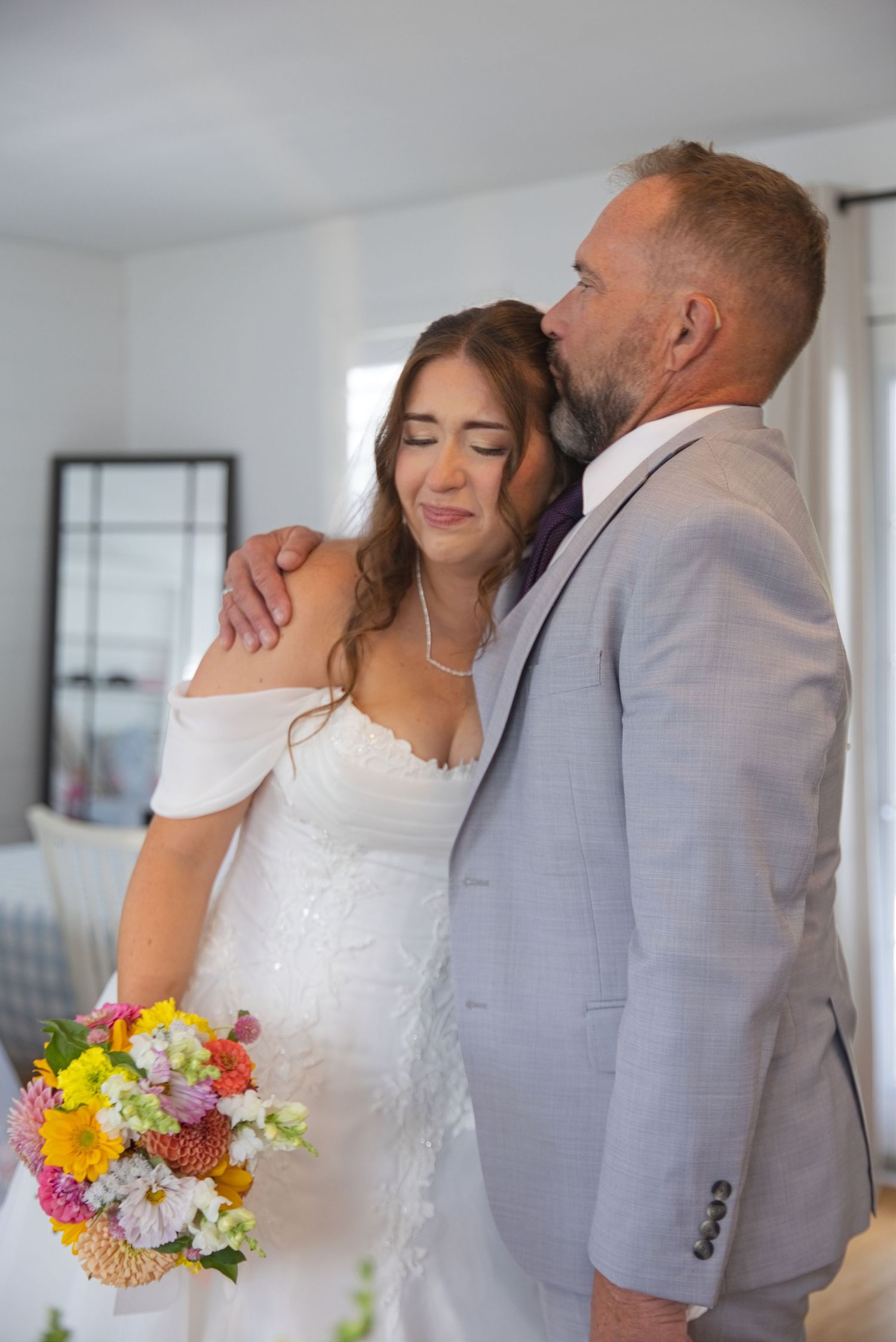 Bride in white dress holding colorful bouquet embraced by groom in gray suit during indoor wedding ceremony.
