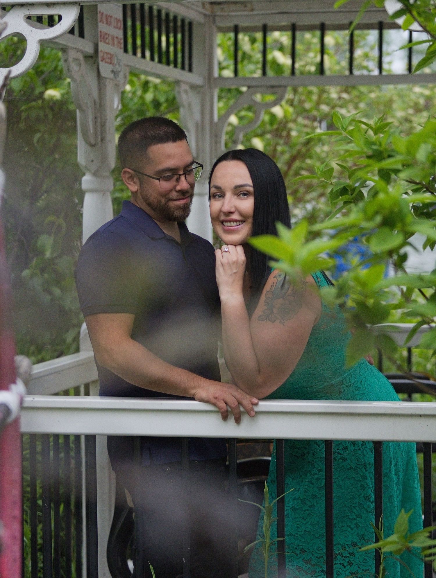 Couple smiling together on a white gazebo porch surrounded by lush green foliage during an outdoor engagement photo session.